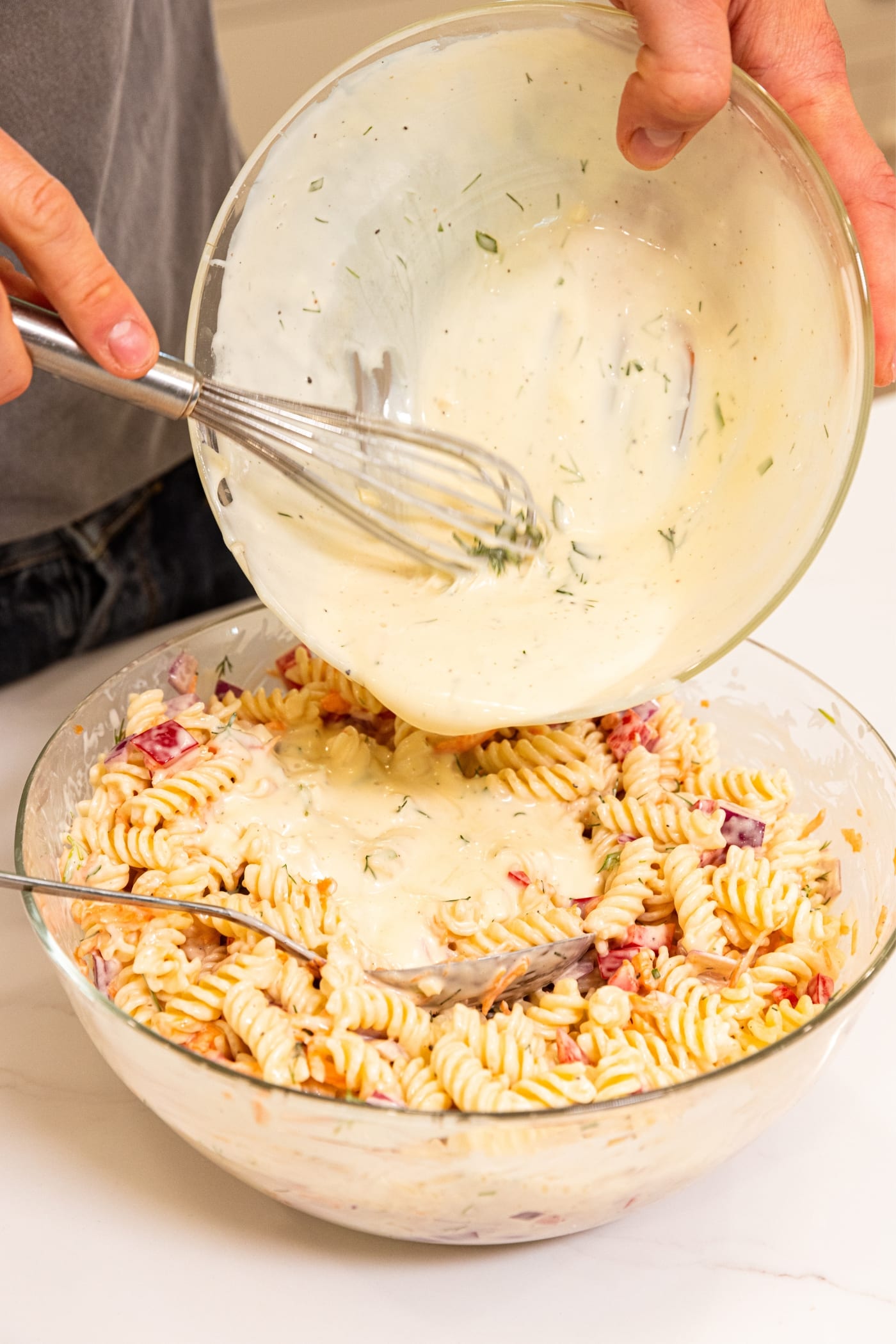 Hand holding glass bowl pouring creamy dressing over the top of pasta salad sitting in another glass bowl on white surface.