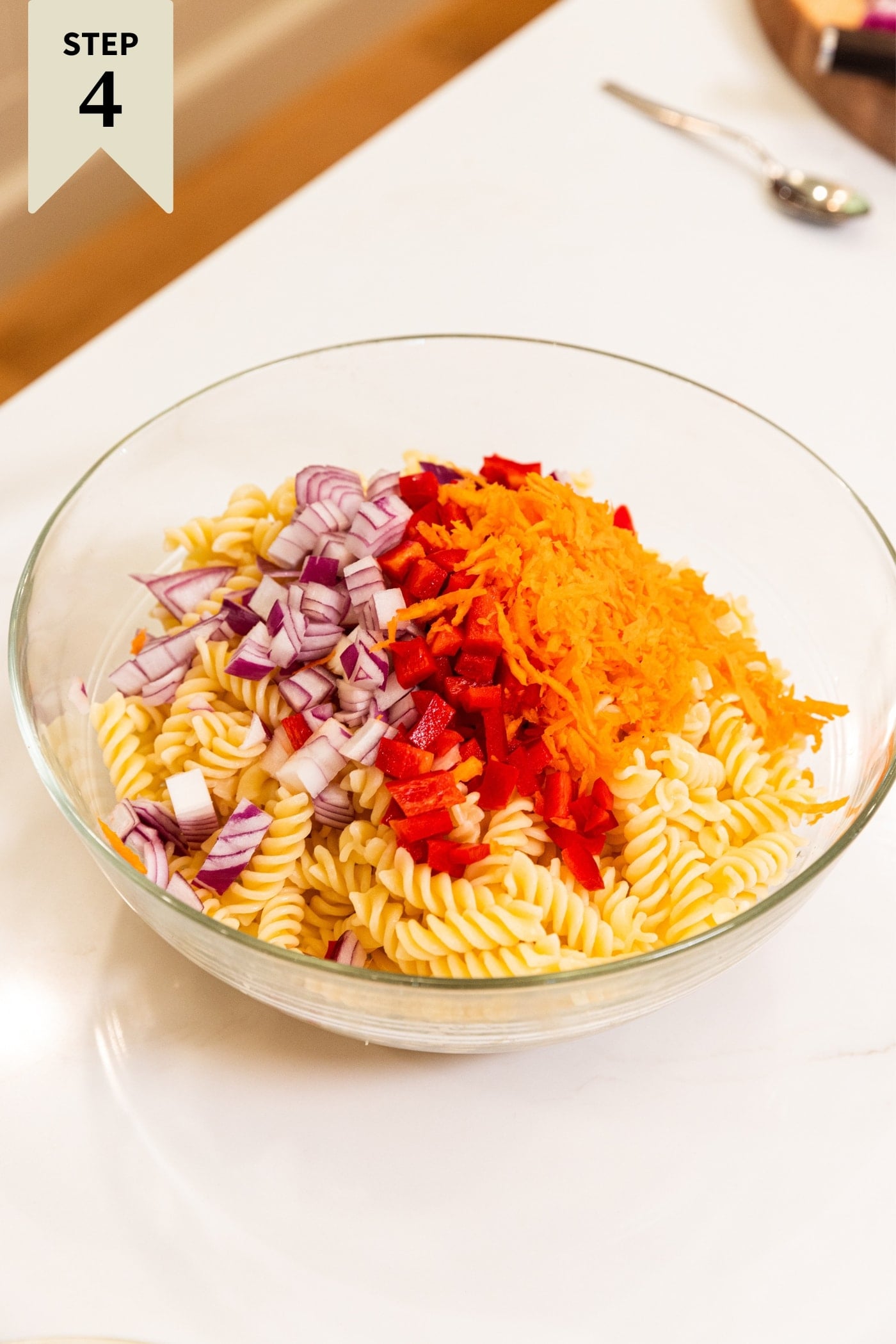 Large glass bowl filled with cooked pasta and topped with diced red onion, diced red bell pepper, and grated carrot all on white surface.