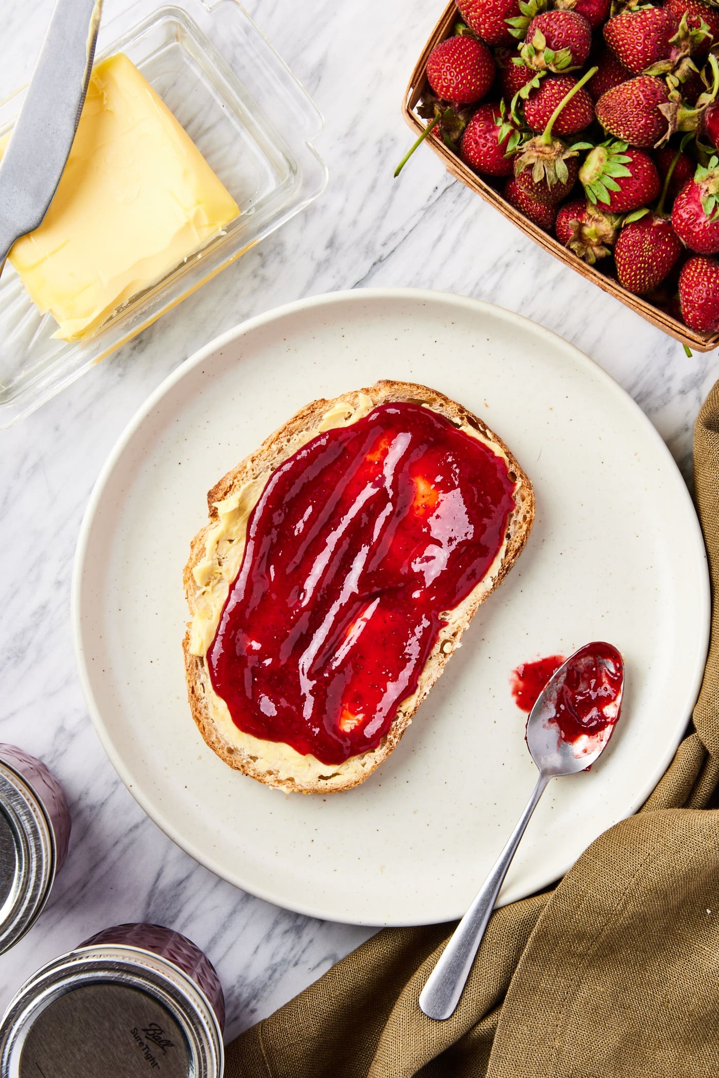 Tan colored plate with slice of bread with butter and strawberry jam slathered on top all on marble surface with more jam jars to the side.