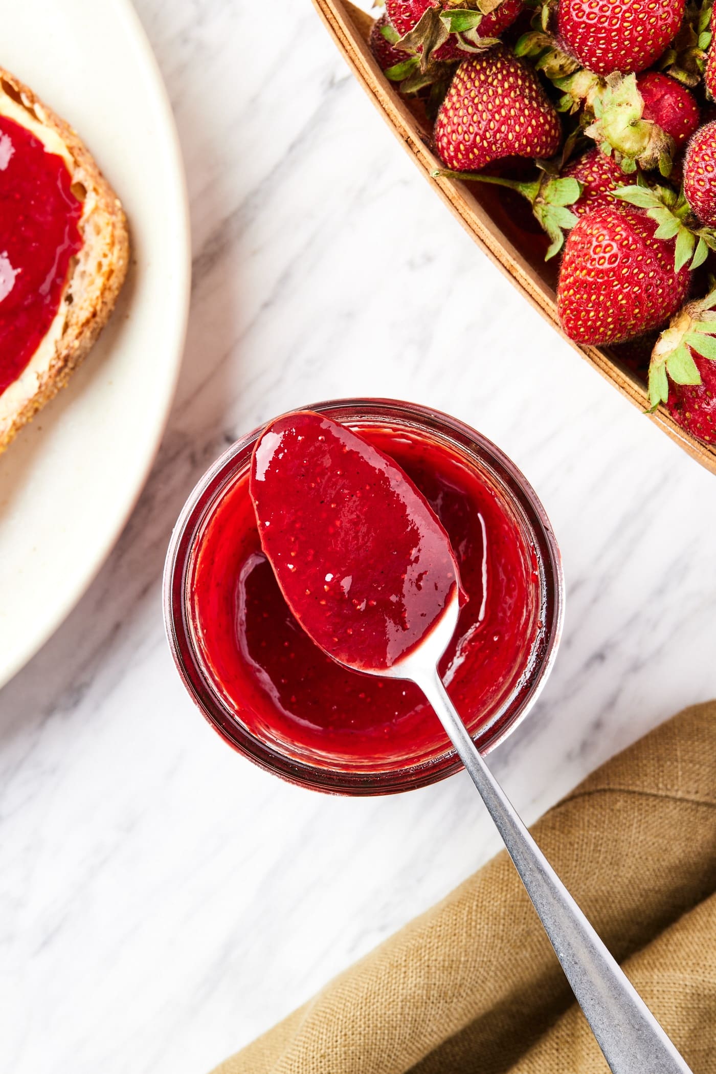 Open glass jam jar with spoon resting on rim with strawberry jam about to drip off with more strawberries to the side.