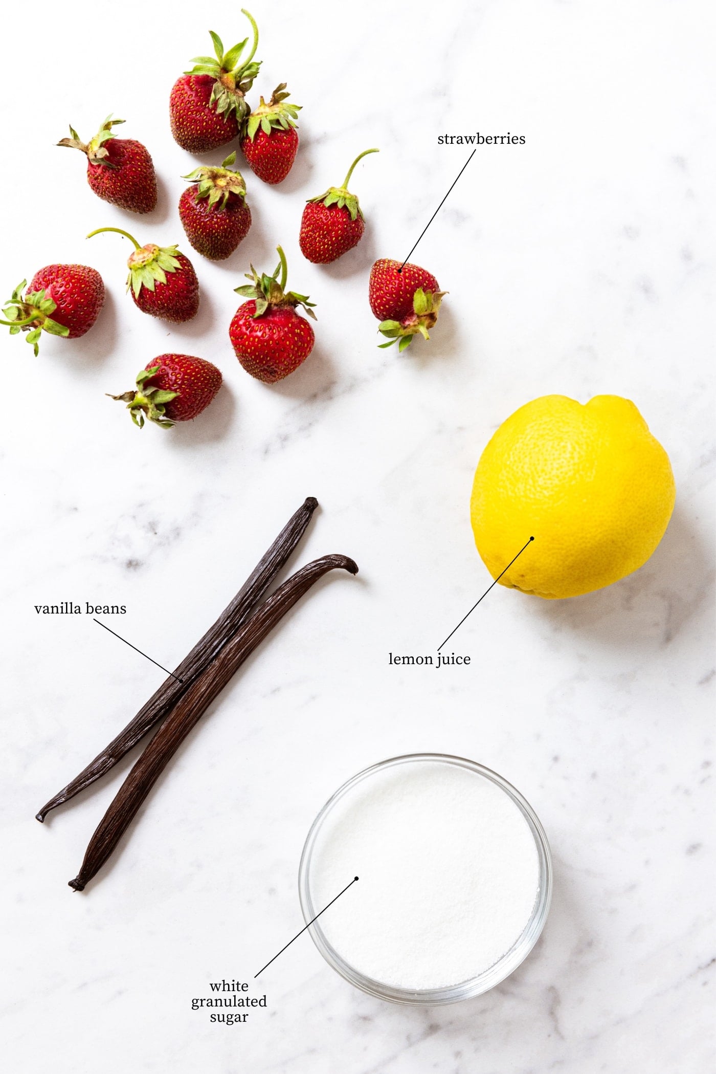 White marble surface filled with four ingredients needed to make strawberry butter including strawberries, lemon, white sugar, and vanilla beans.