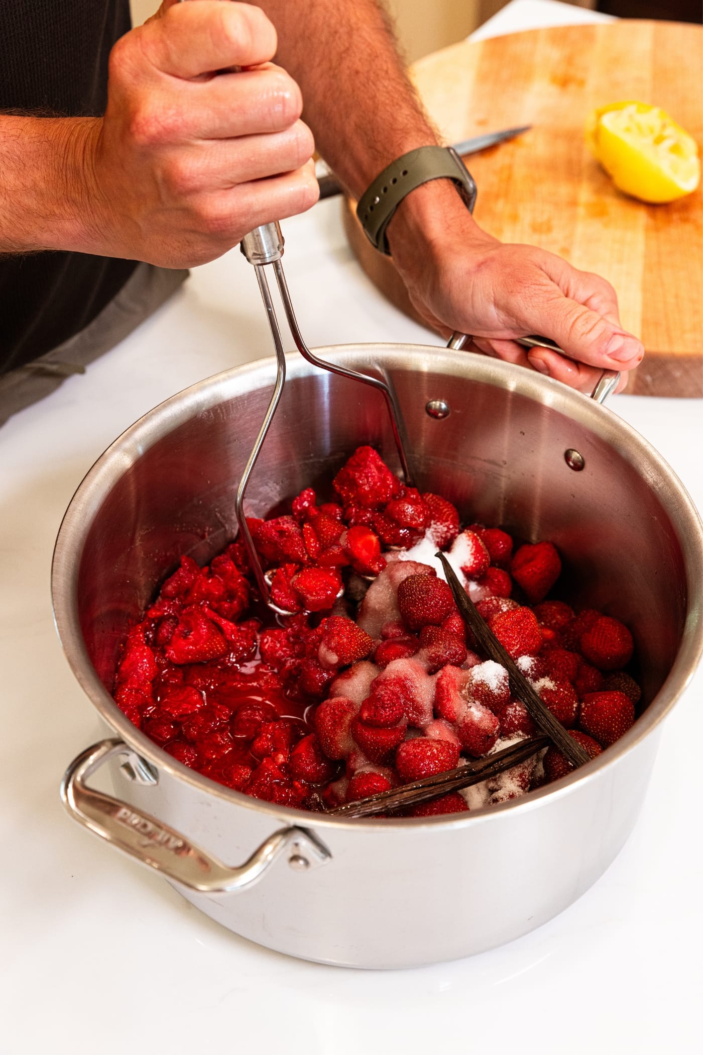 Large metal stockpot filled with strawberries with hand using masher to mash the strawberries with cutting board in background.