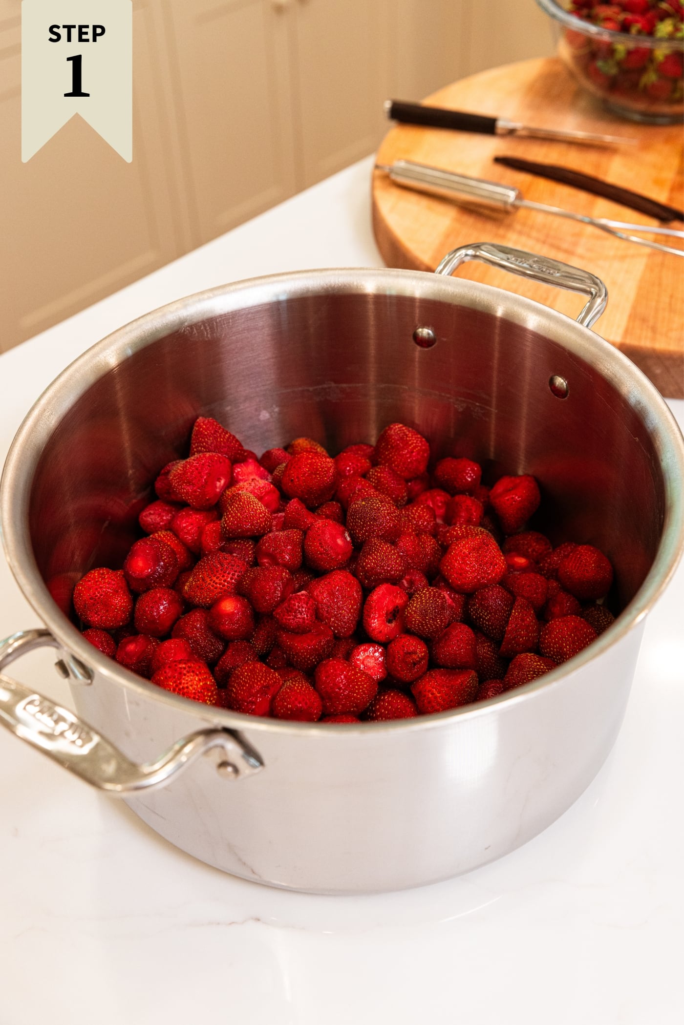 Large metal stockpot filled with whole hulled strawberries on white surface with wood cutting board in background.