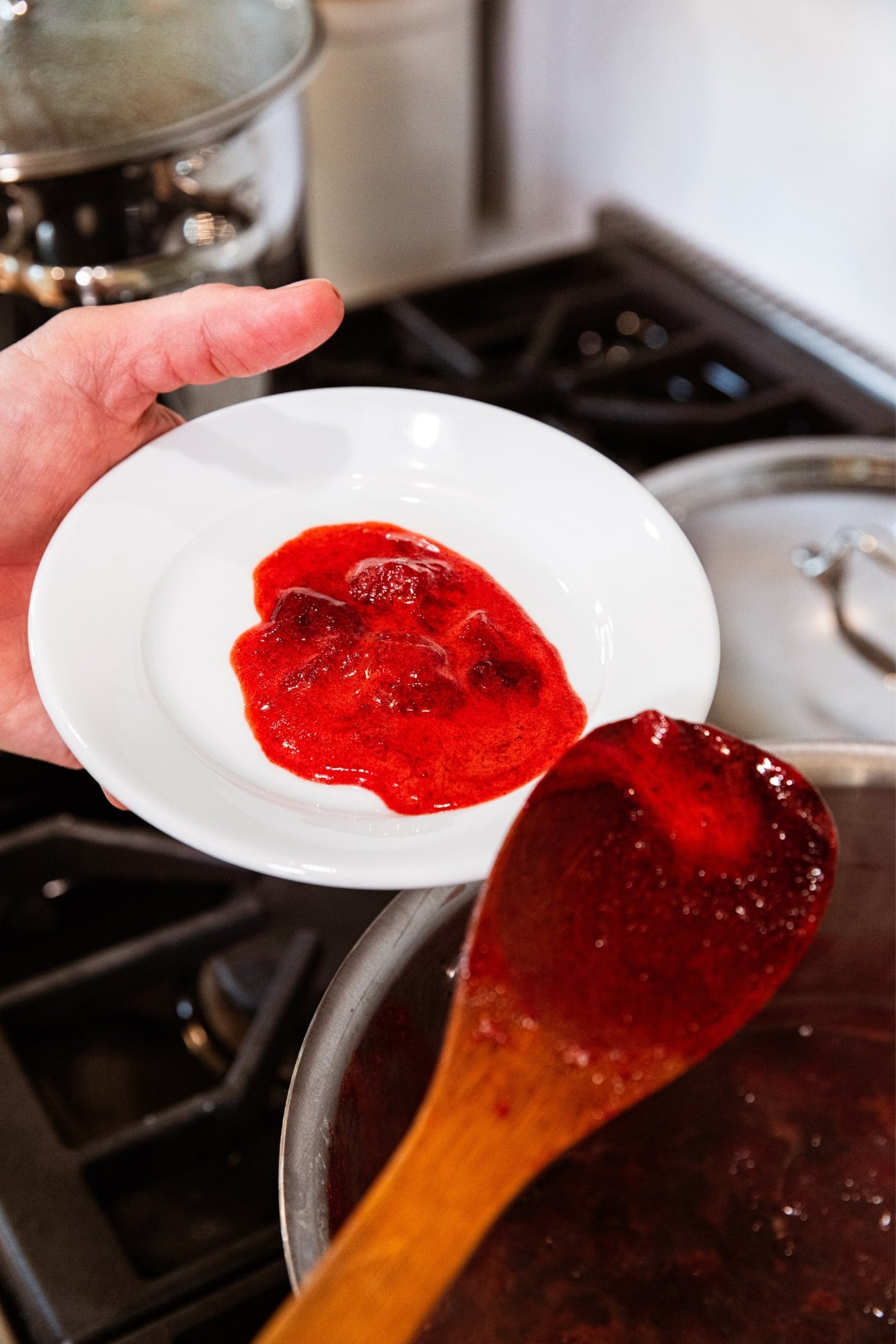 Hand holding white plate with red colored foam mixture from strawberry butter mixture with wooden spoon spooning it out of pot.
