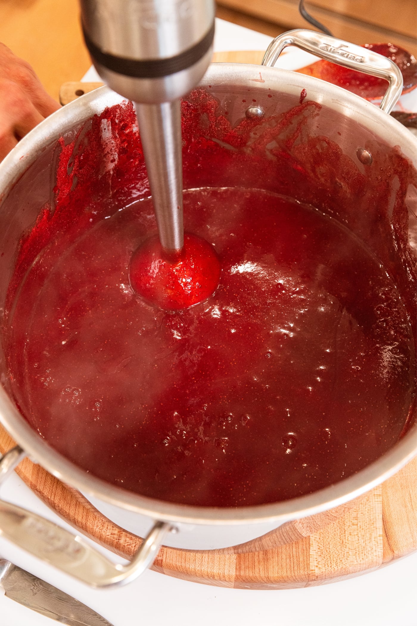 Immersion blender resting in large metal pot filled with strawberry butter with smooth texture.