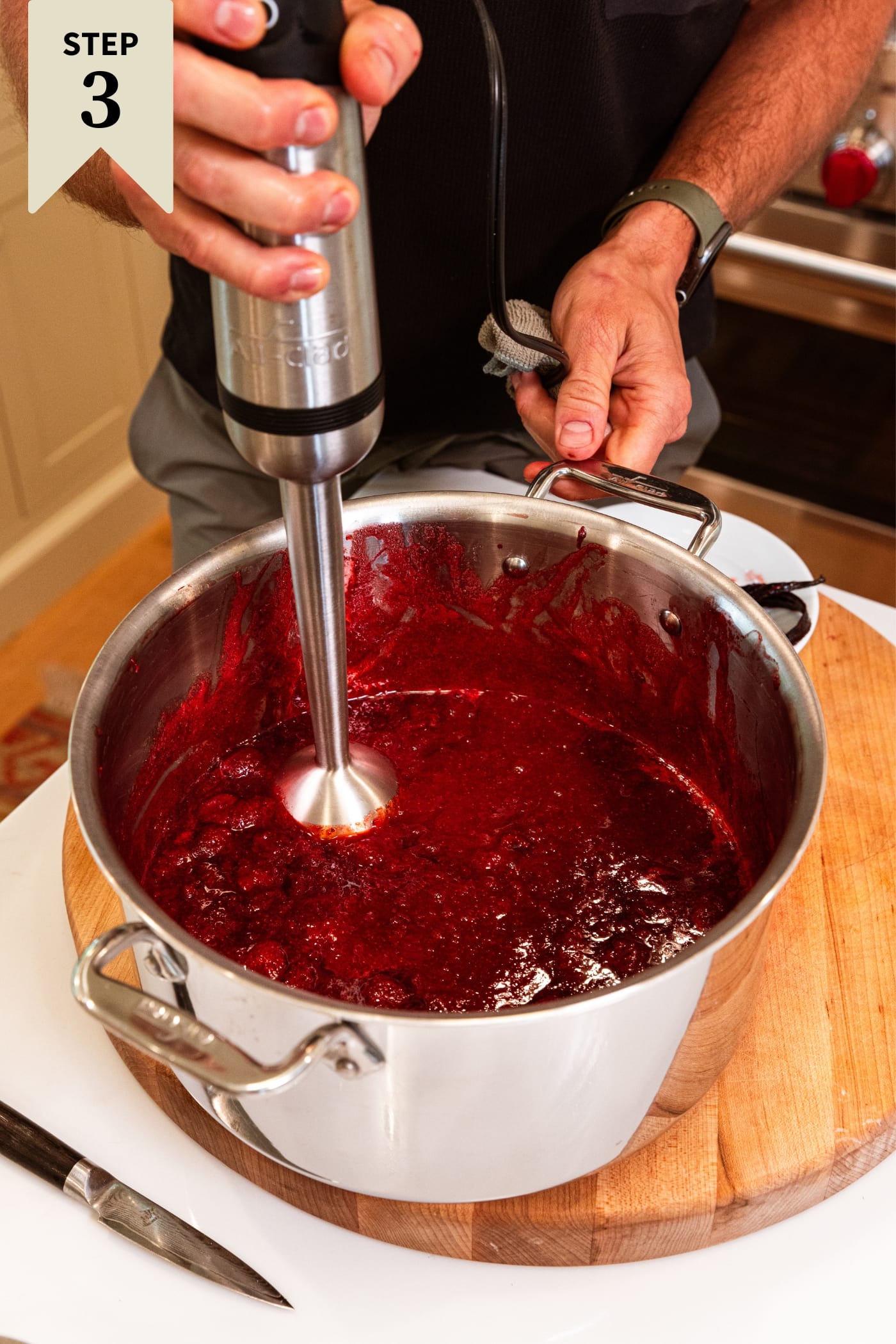 Hand holding immersion blender blending together strawberry and sugar jam mixture in large metal pot sitting on wooden cutting board.
