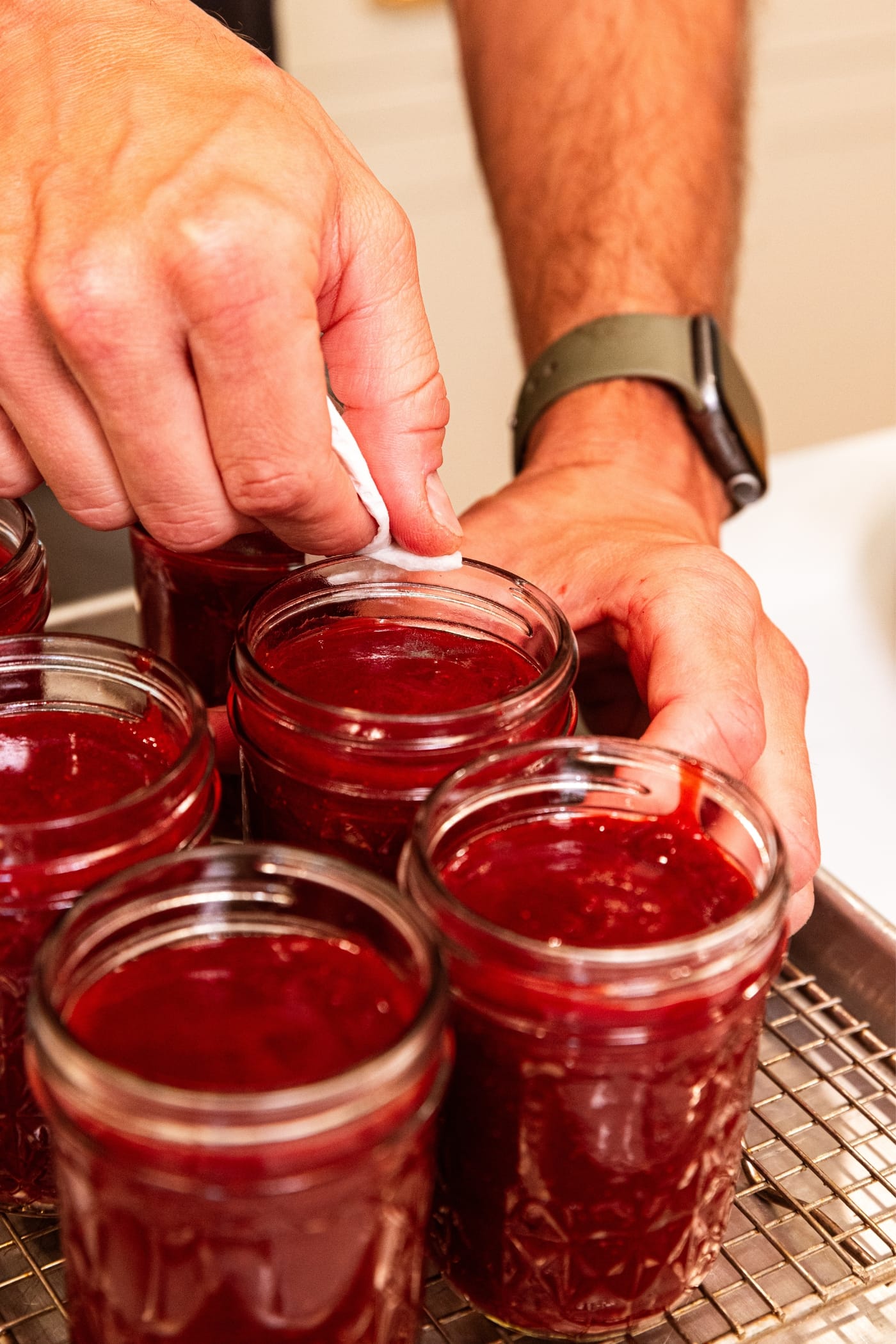 Hand using piece of paper towel to wipe off the rim of a glass jam jar filled with strawberry butter.