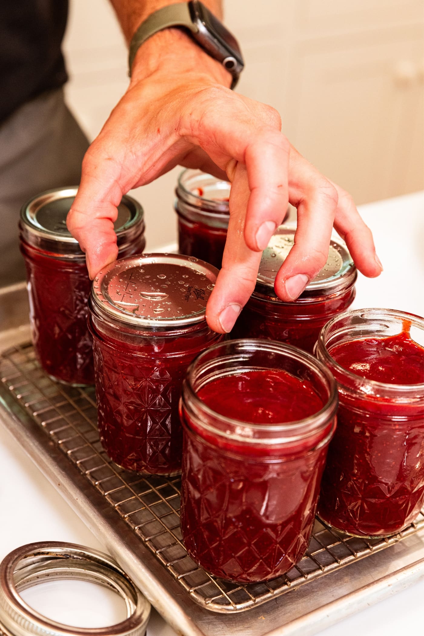 Hand placing lid on top of glass jam jar filled with strawberry butter with all jars resting on metal cooling rack.