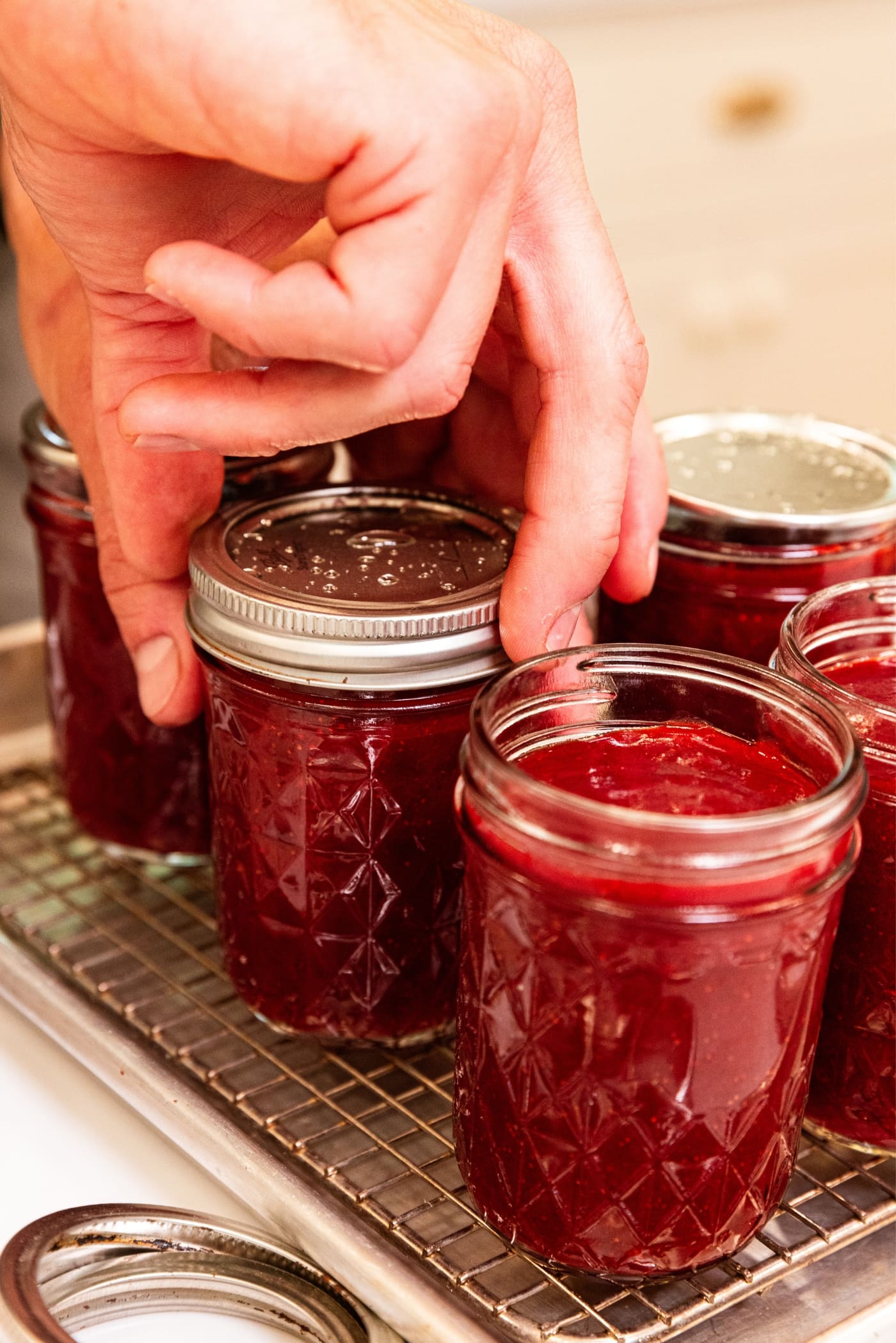 Hand turning ring on top of glass jam jar filled with strawberry butter with lid also on top resting on metal cooling rack.