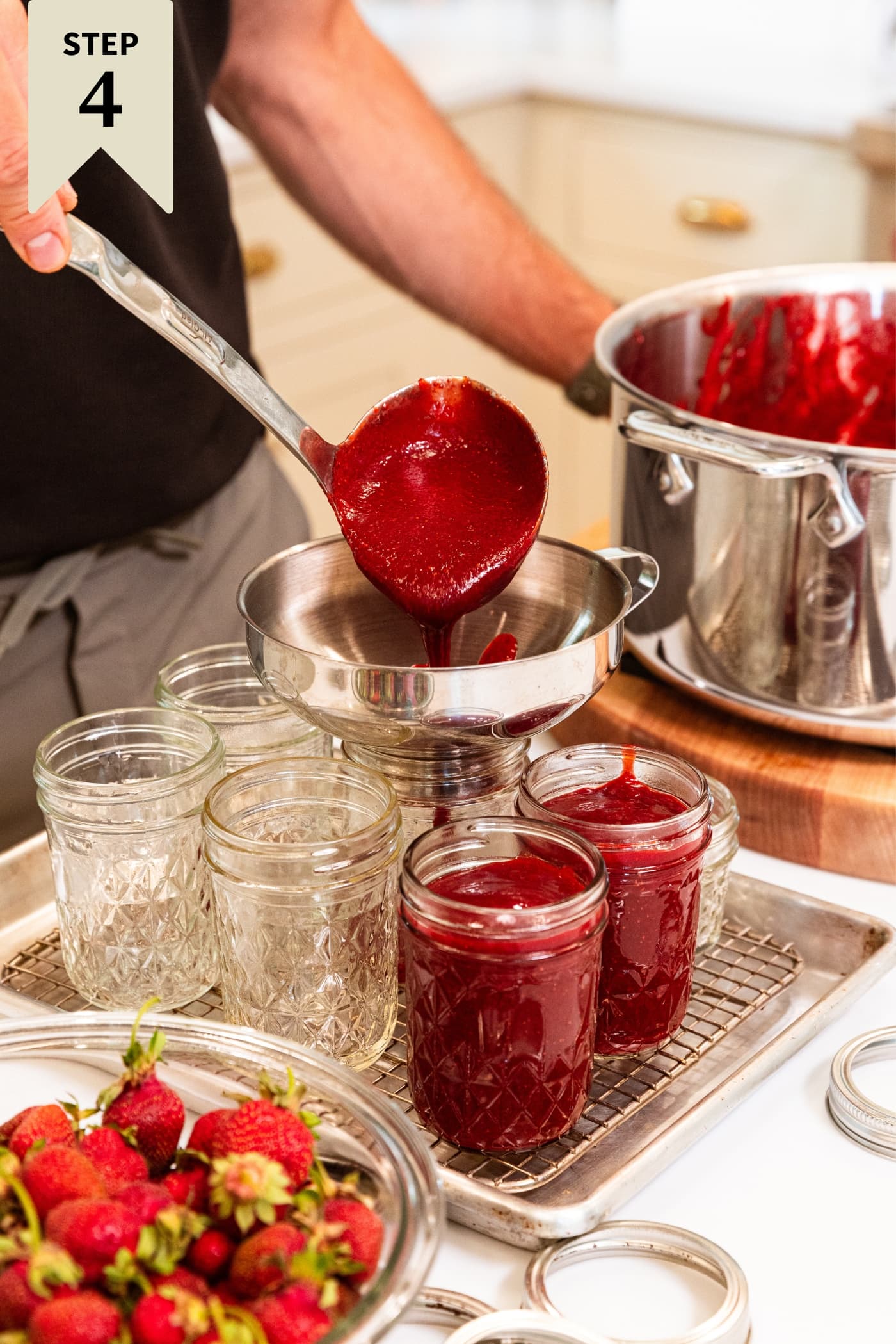Hand using ladle to pour strawberry butter into glass jam jars using a funnel with large metal stockpot in the background.