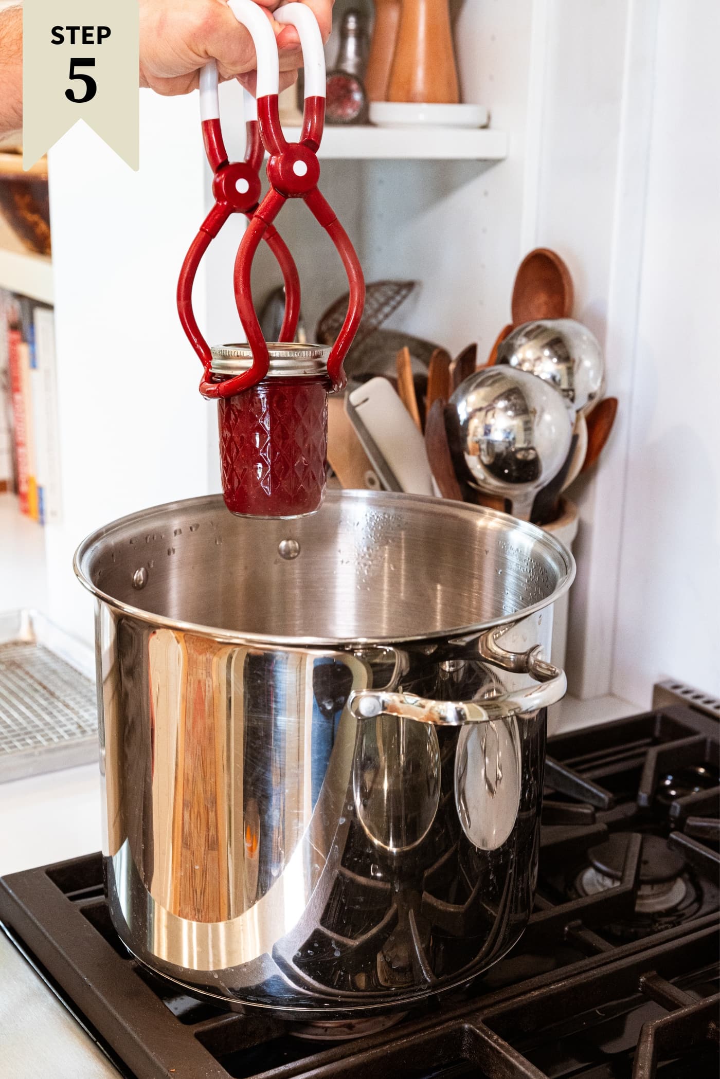 Large metal canning pot resting on stovetop with hand holding jar lifter placing jam jar filled with strawberry butter into the pot.