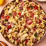 Large brown bowl filled with Italian tortellini pasta salad with red onion, bell pepper, artichoke hearts, parsley, olives, and more all on taupe surface.