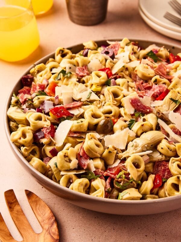 Large brown bowl filled with Italian tortellini pasta salad sitting on taupe surface filled with lots of additions with lemonade in background.