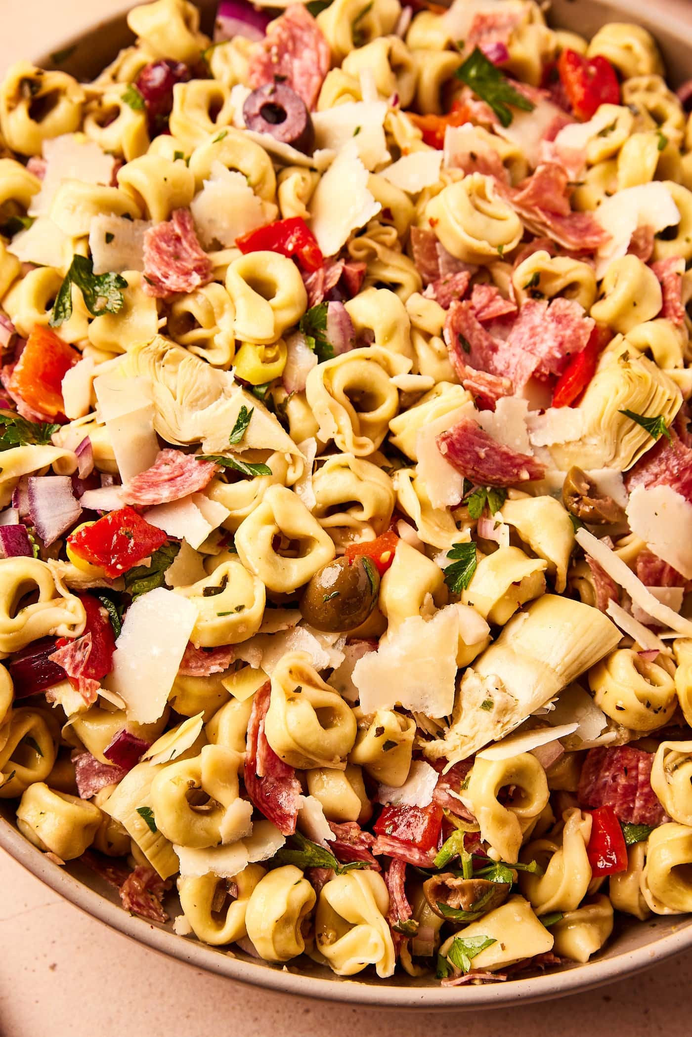 Cooked tortellini pasta in large bowl tossed with pieces of red bell pepper, artichoke hearts, sliced olives, red onion, Parmesan cheese, and more.