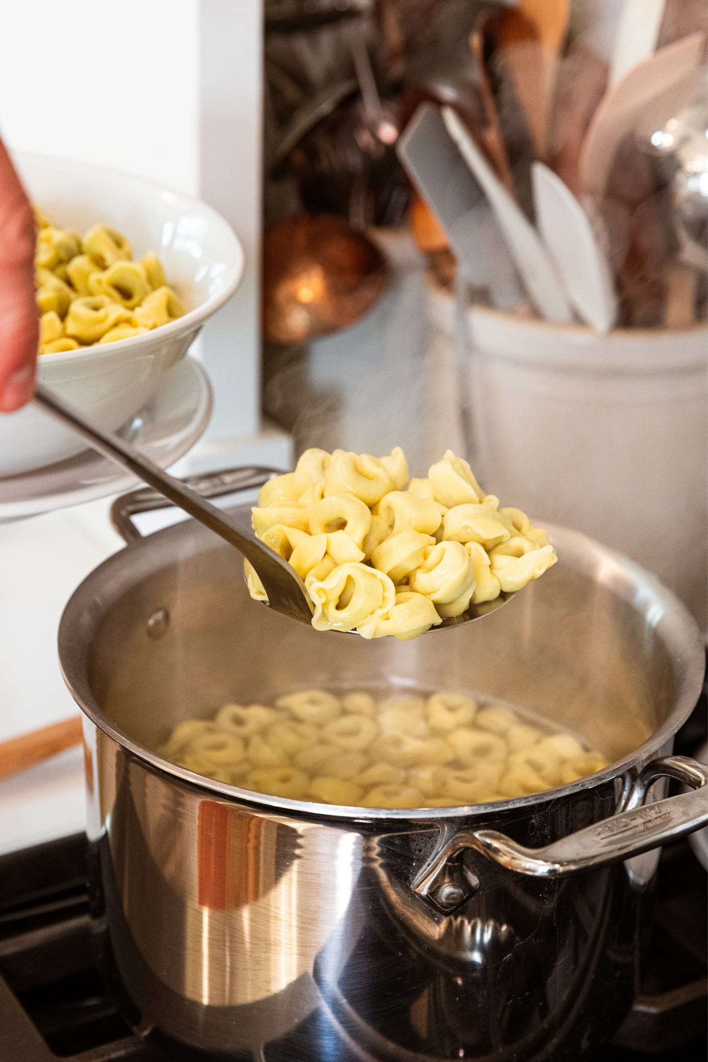 Hand holding ladle pulling cooked stuffed tortellini pasta out of pasta water in large stockpot on stovetop.