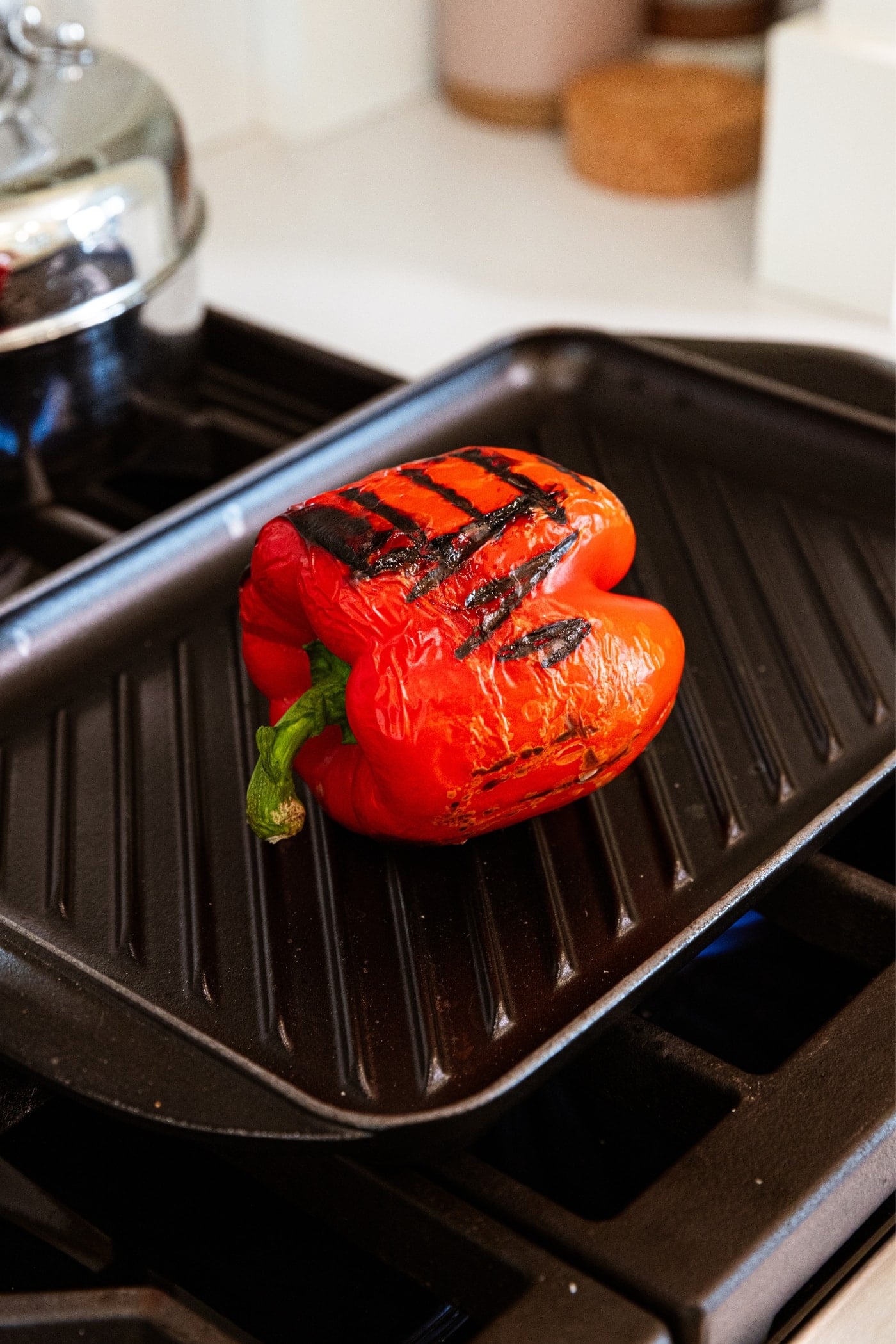 Blackened red bell pepper sitting on grill pan on stovetop with grill marks all over the exterior skin.