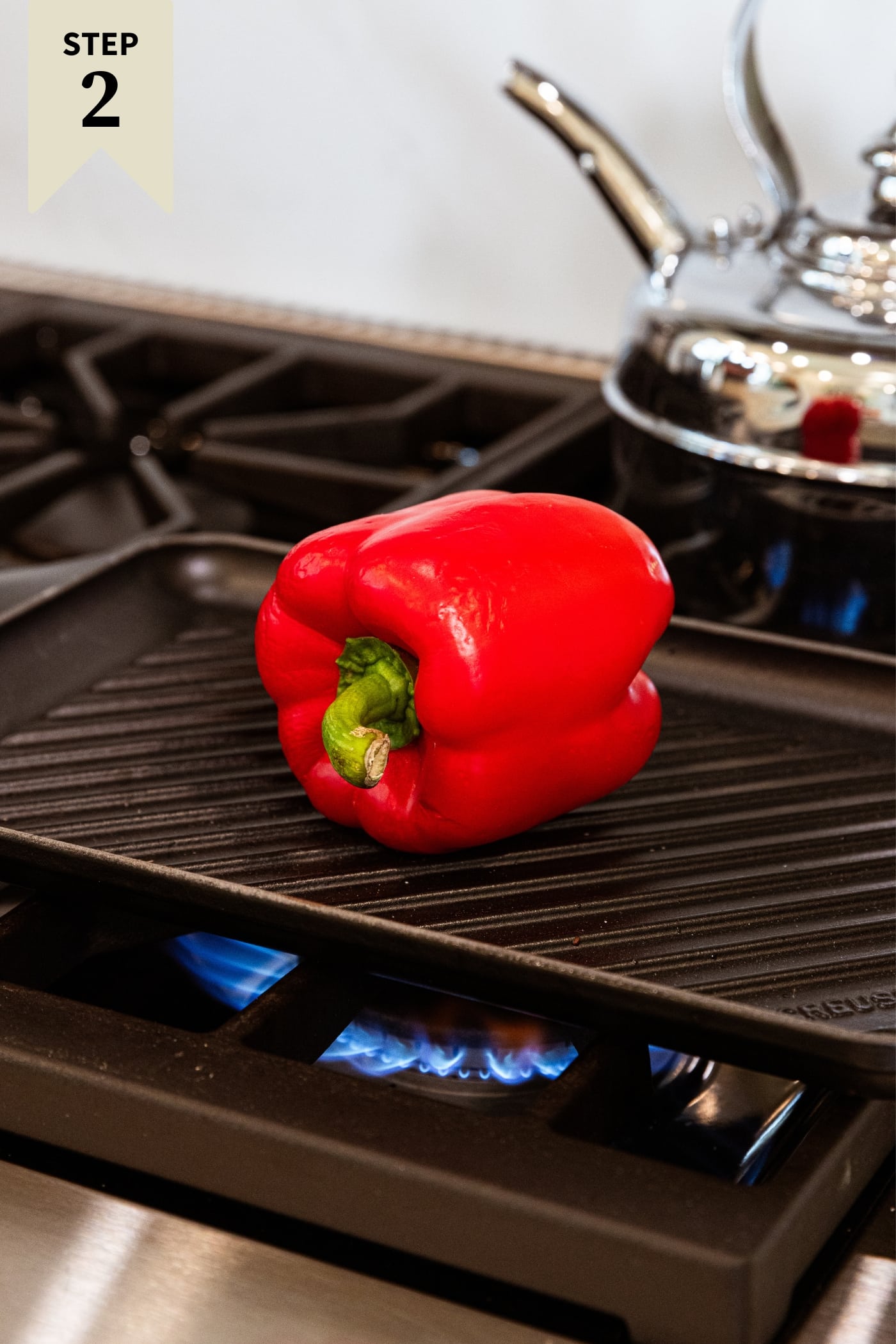 Red bell pepper sitting on grill pan over flame on stovetop before becoming blackened.