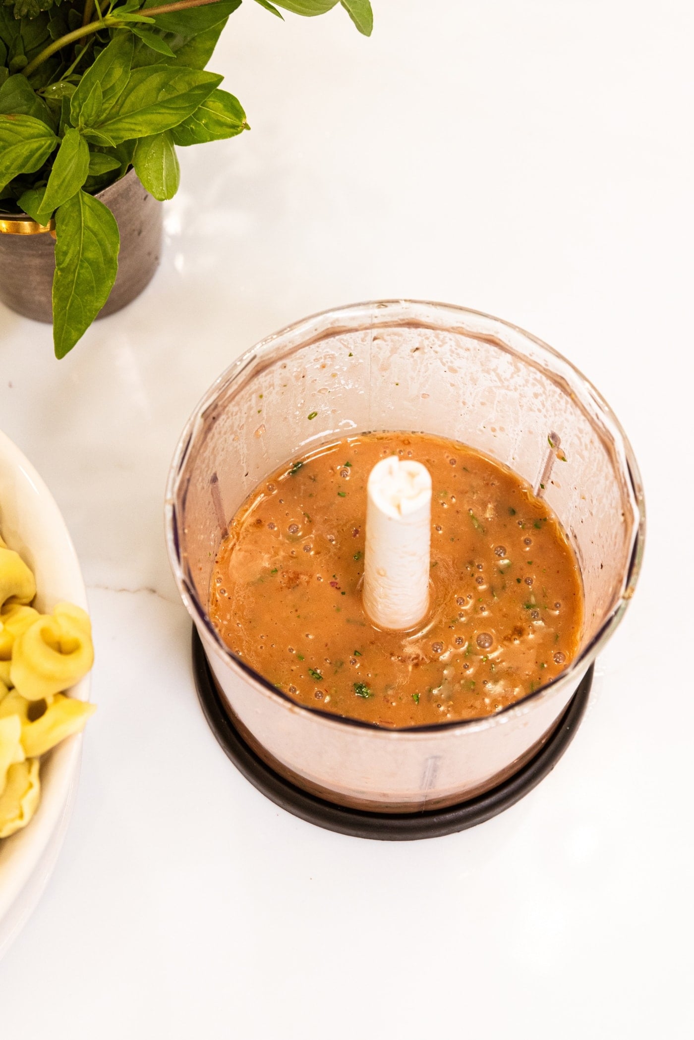 Container of a small food processor with mixed Italian dressing in container with bowl of pasta to the side.