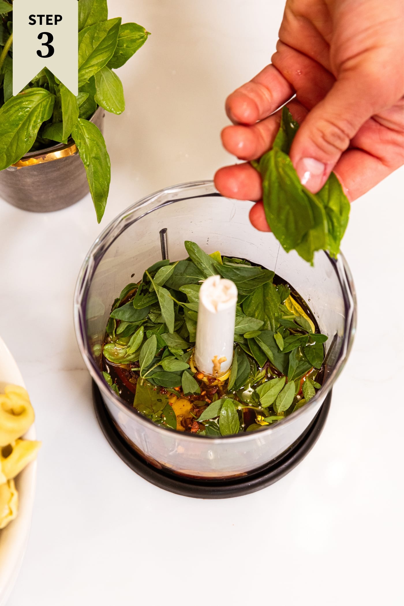 Hand holding basil leaves about to throw into the container of a food processor already filled with herbs and oils.