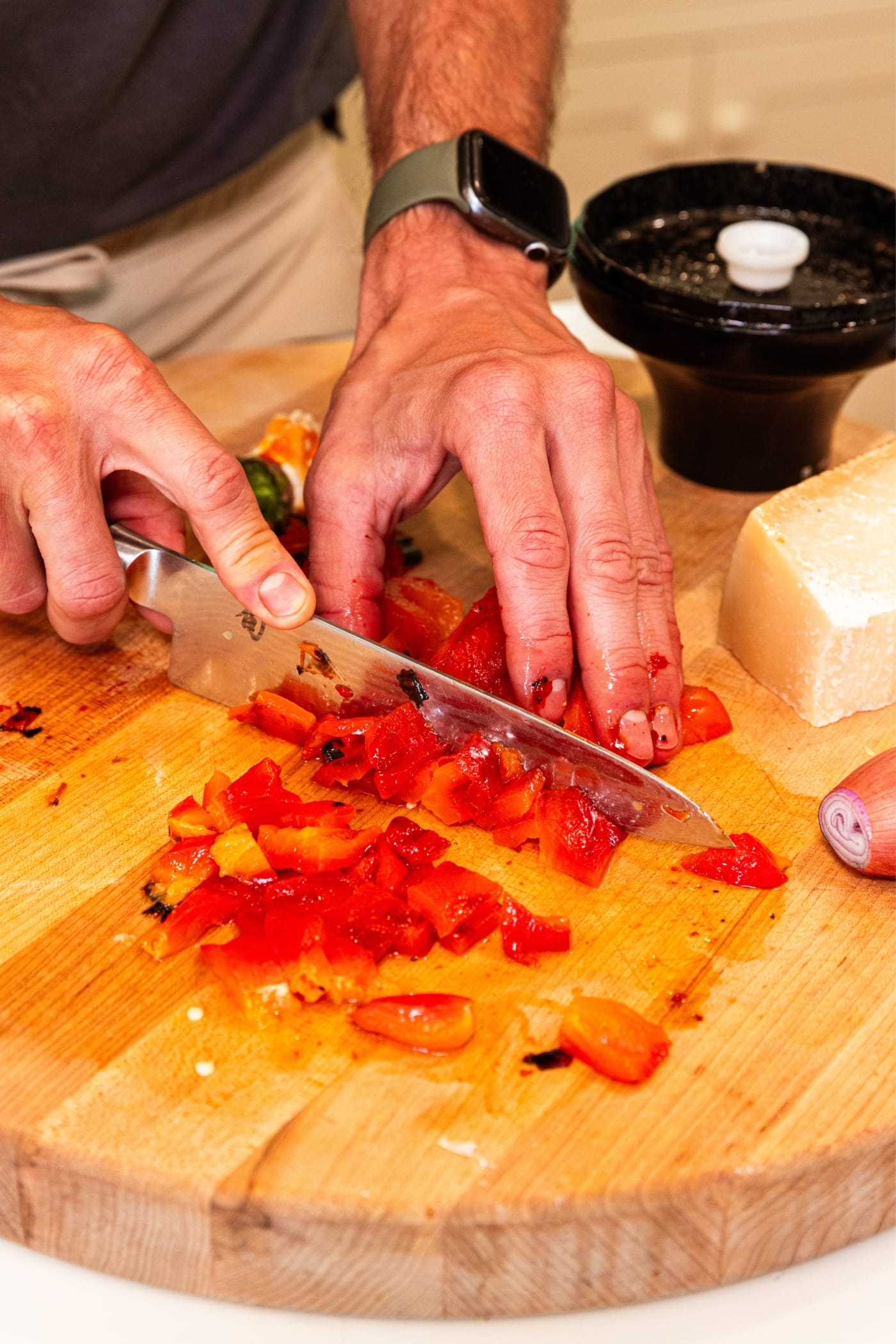 Hands using a knife to dice grilled red bell pepper on wooden cutting board.