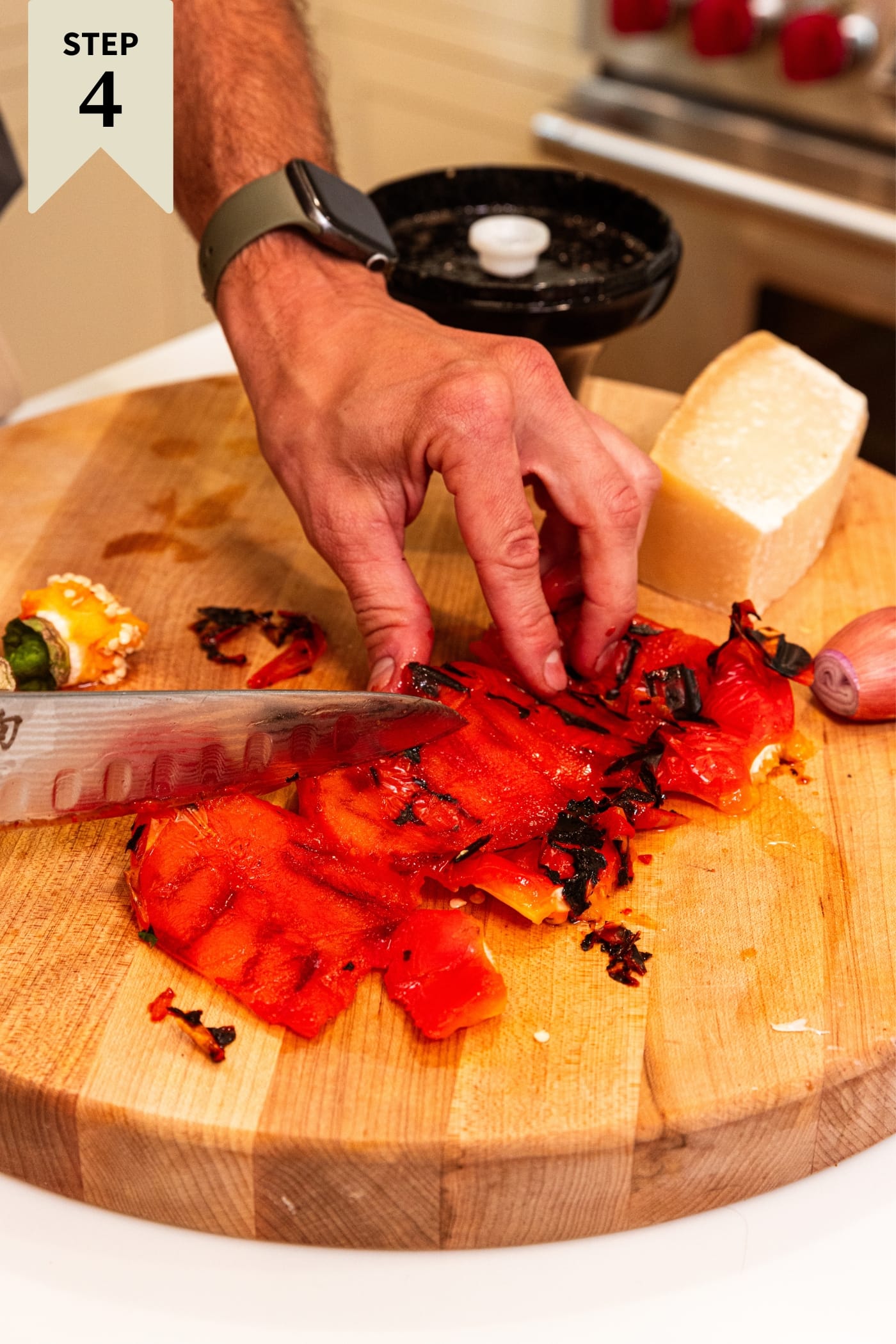 Hand using a knife to scrape the blackened skin from a red bell pepper on a wooden cutting board.