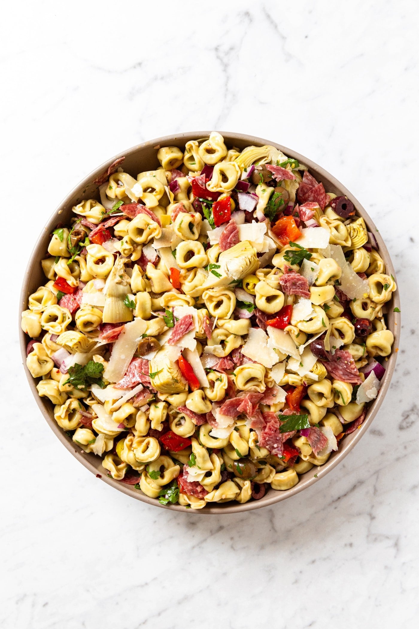 Large brown bowl filled with cooked tortellini pasta tossed with other ingredients including olives, Parmesan cheese, bell pepper, and more.