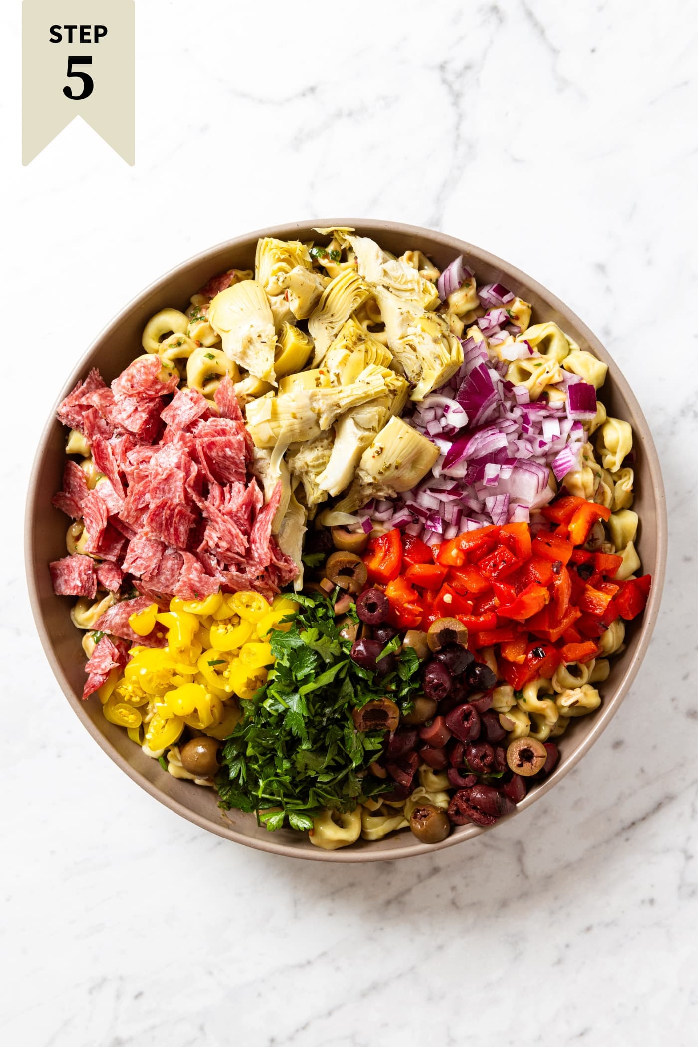 Large brown bowl sitting on white marble surface filled with cooked tortellini and lots of toppings including olives, artichoke hearts, parsley, salami, and more.