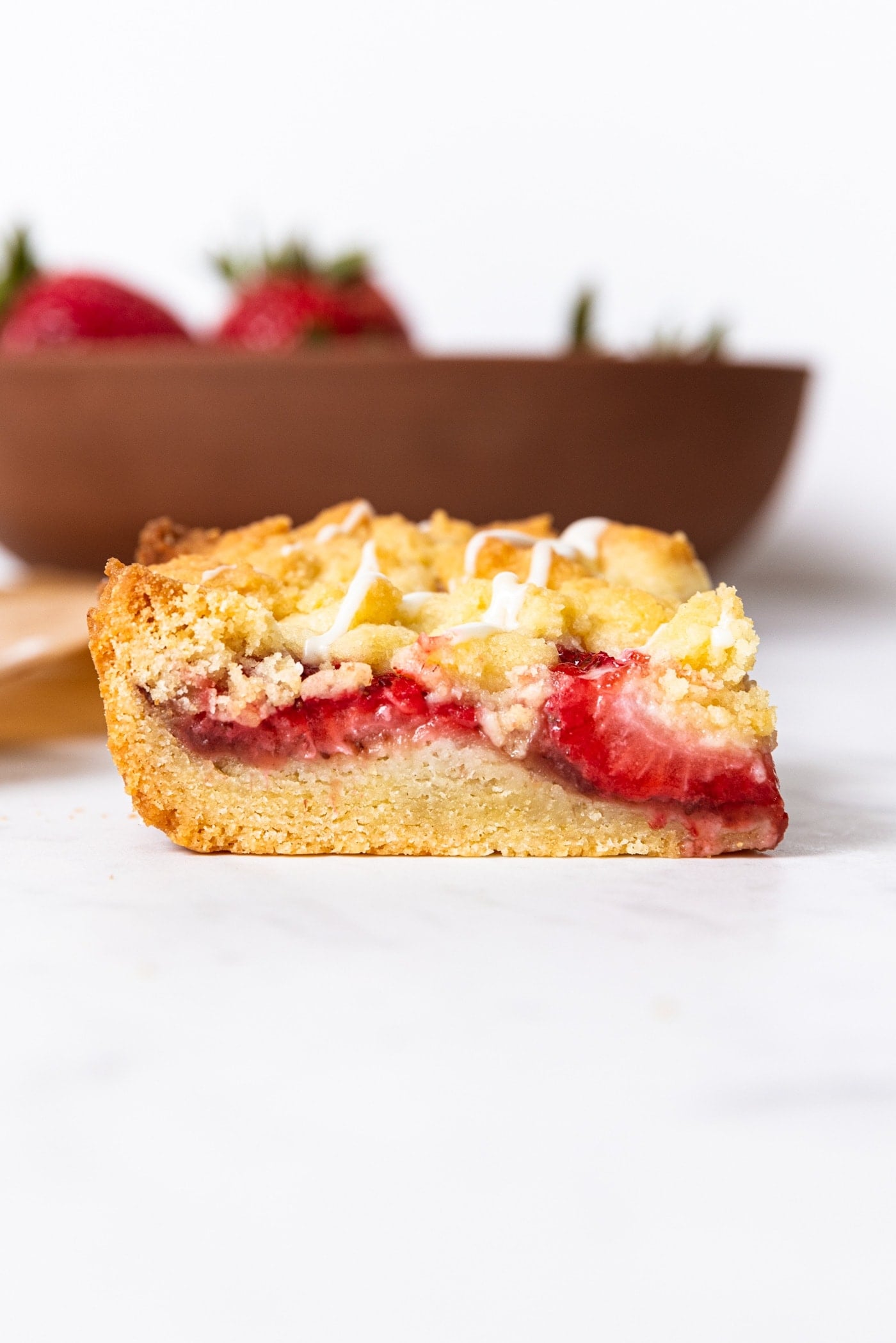 Strawberry crumb bar sitting on white marble surface with bowl of strawberries in the background showing interior texture of the bar.