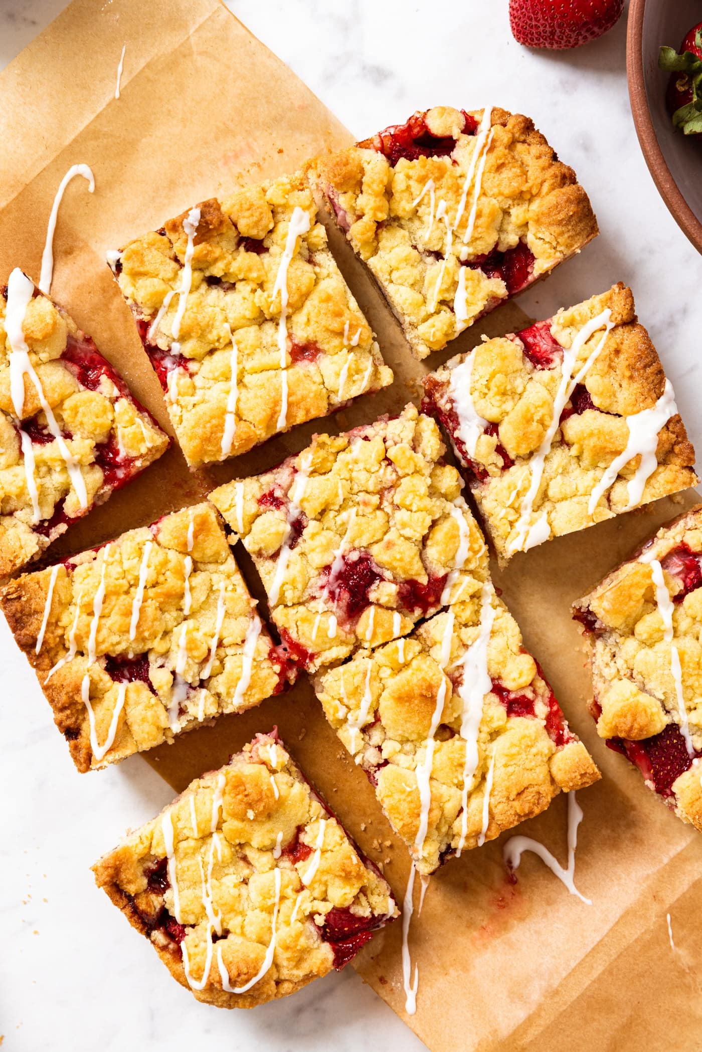 White marble surface with strawberry crumb bars sliced into pieces sitting on top of brown piece of parchment paper with strawberries in bowl.