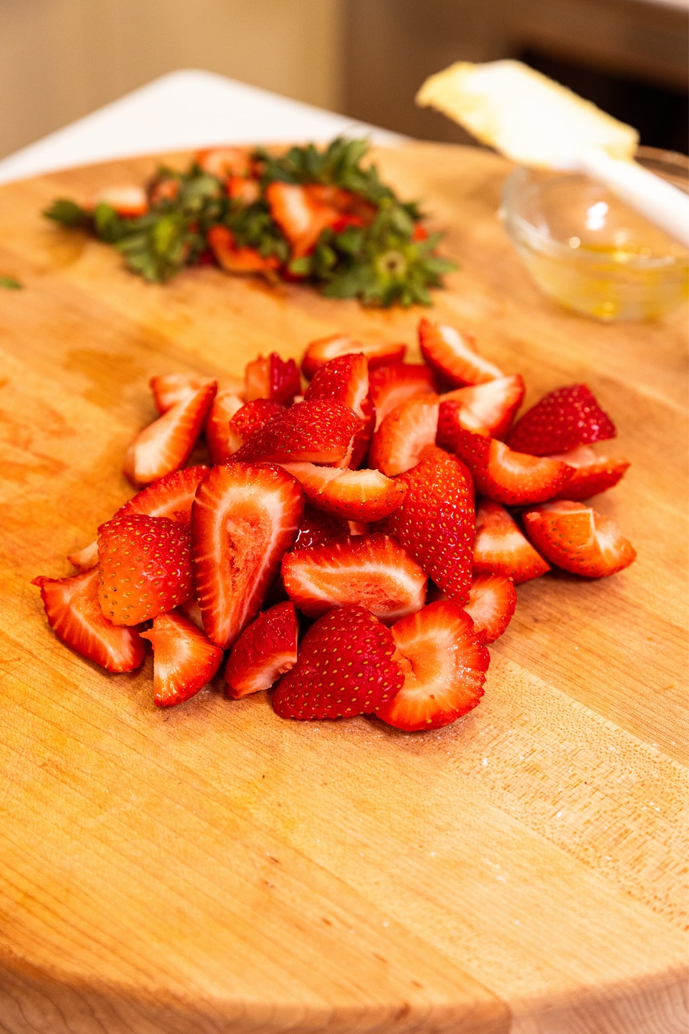 Wood cutting board with sliced and quartered strawberries with tops sitting behind.