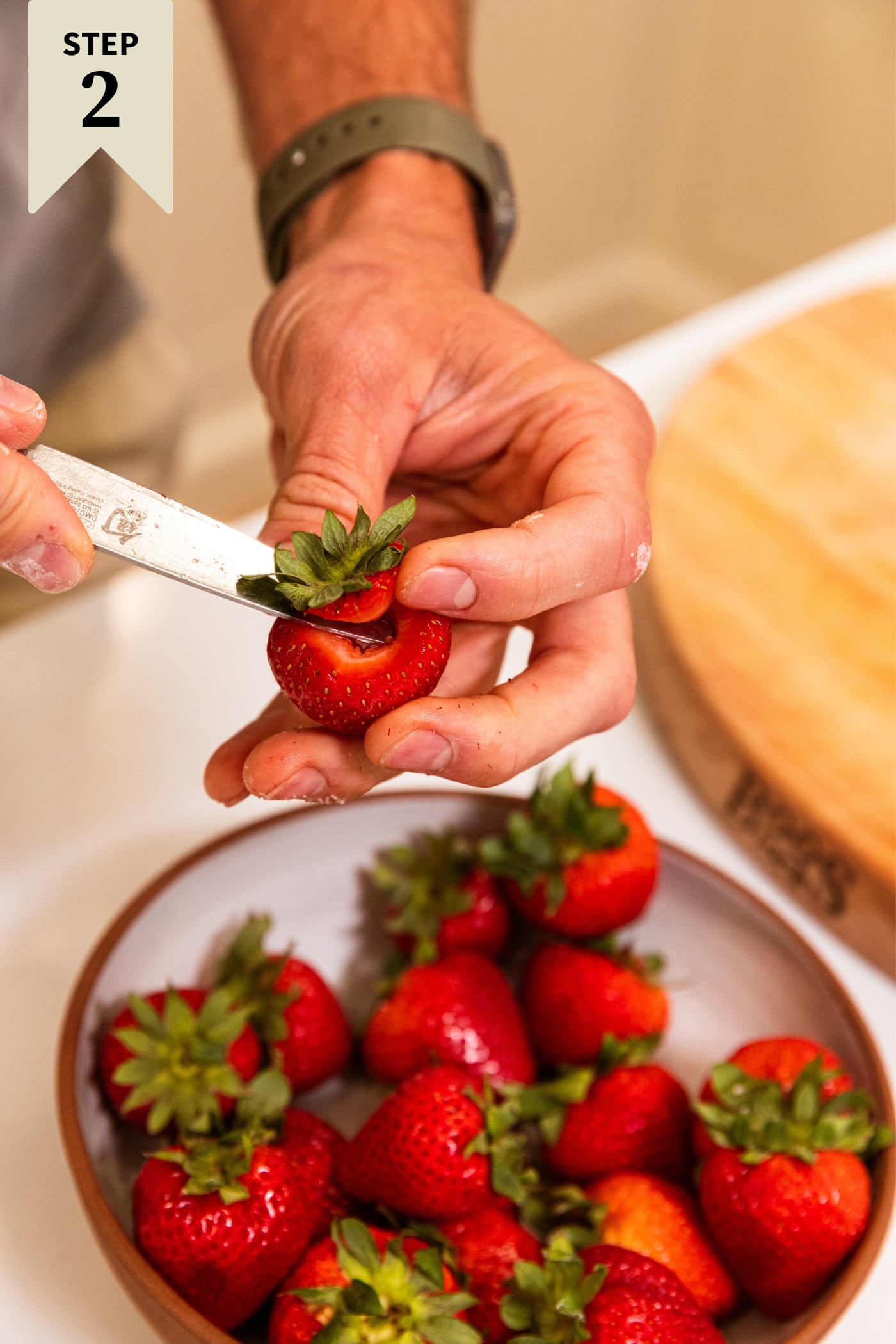Hand holding knife hulling strawberries with more strawberries in a bowl below.