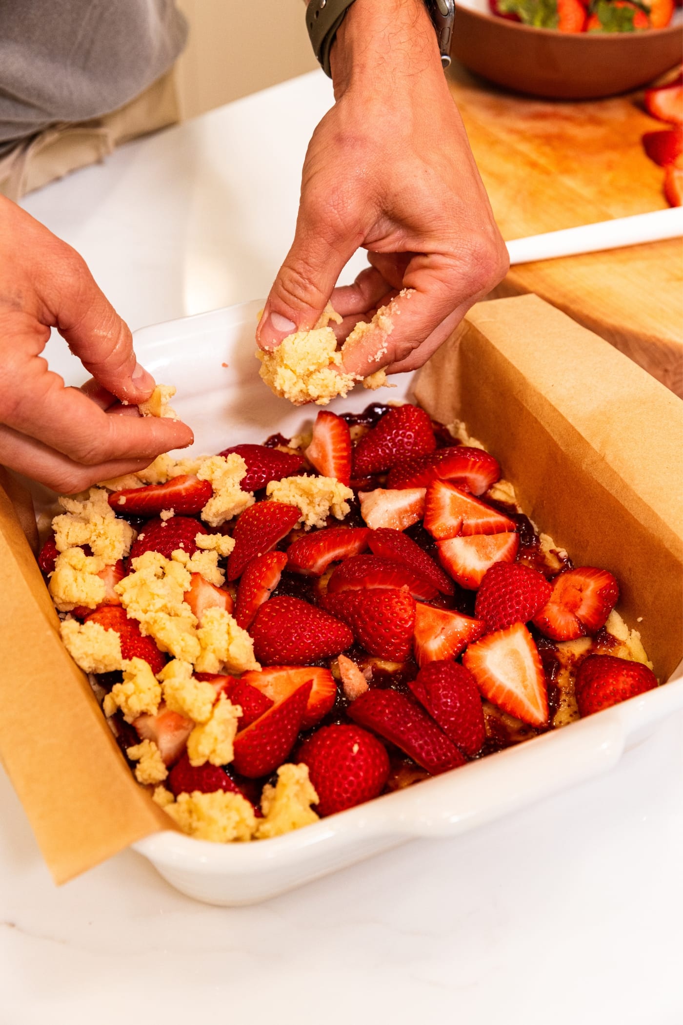 Two hands sprinkling crumbled dough on top of strawberries and jam in a square baking dish with parchment paper.