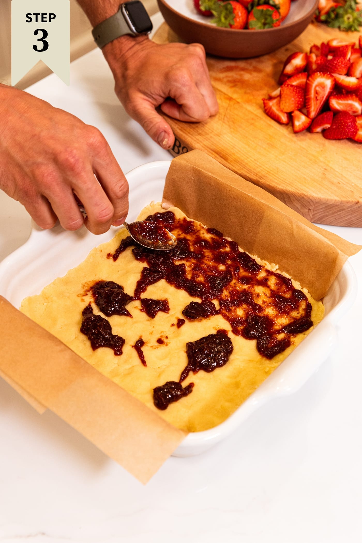 Hand using spoon to smooth out strawberry jam on top of a crust pressed into a square baking dish lined with brown parchment.