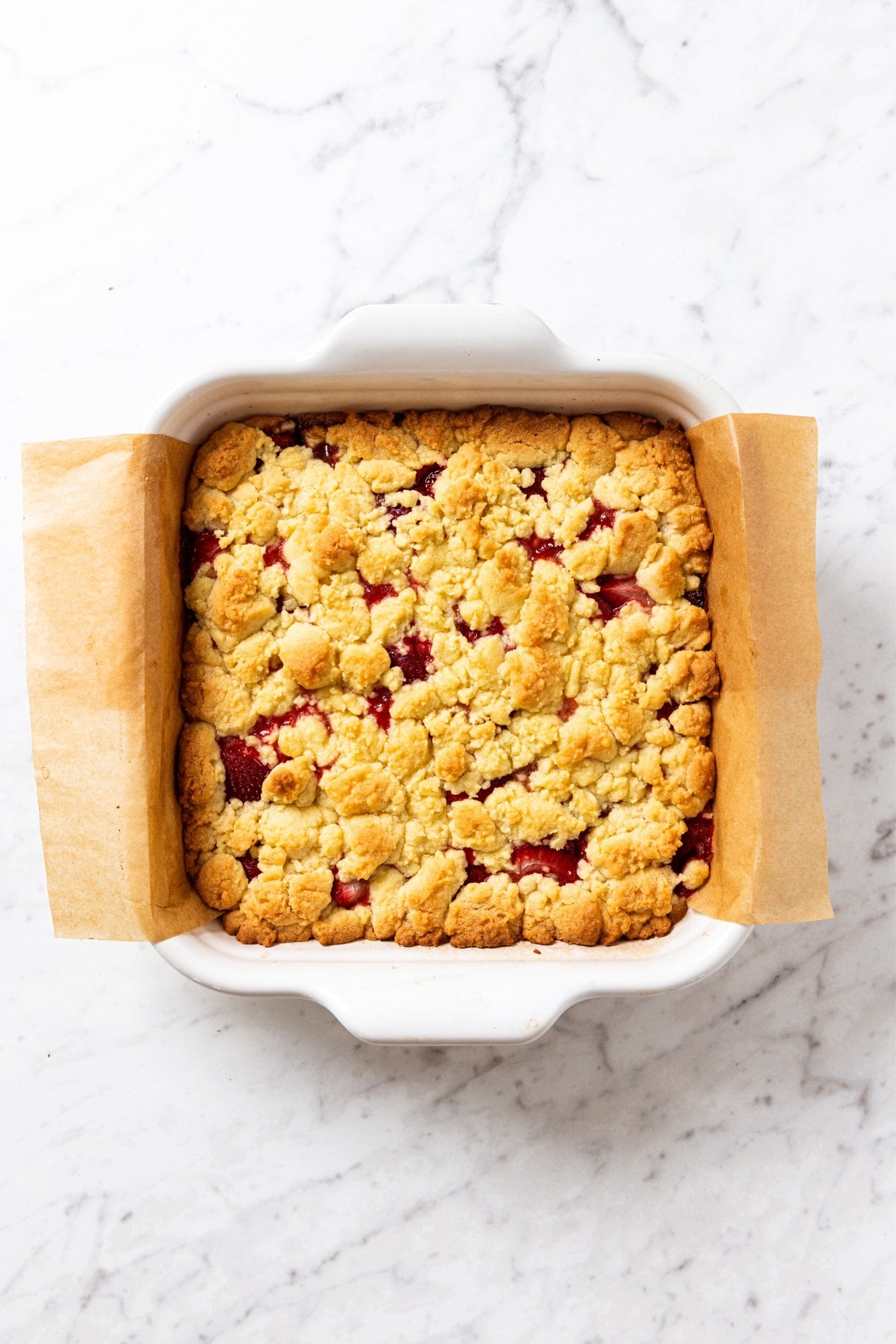 White baking dish sitting on white marble surface with baked strawberry crumb bars right after coming out of the oven.