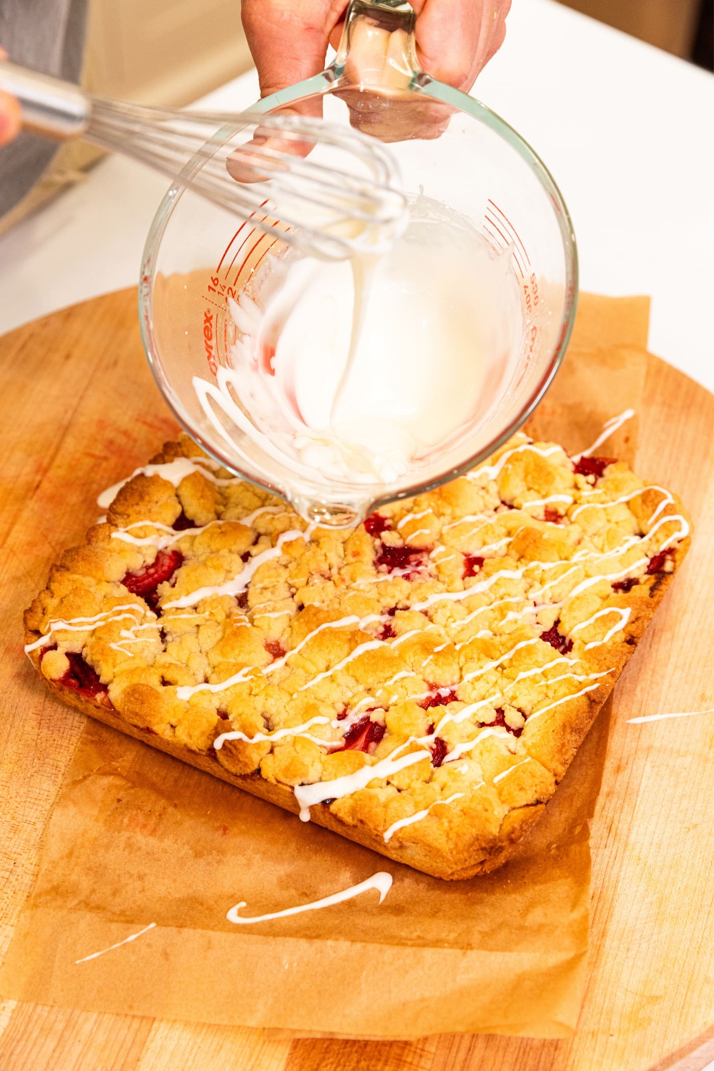 Hand holding glass measuring cup pouring glaze on top of strawberry crumb bars with golden top.