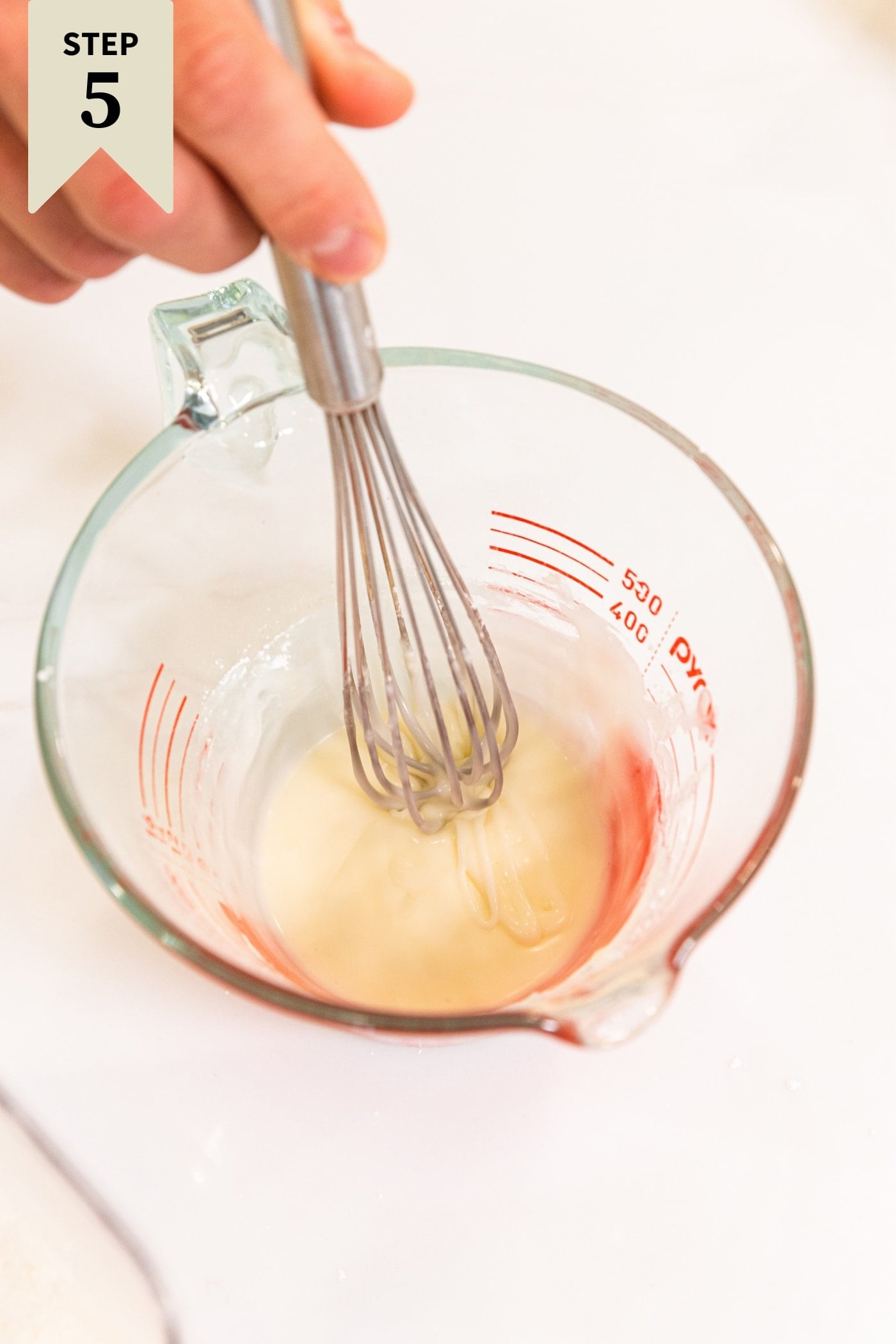 Glass measuring cup with glaze being mixed with whisk on white surface.