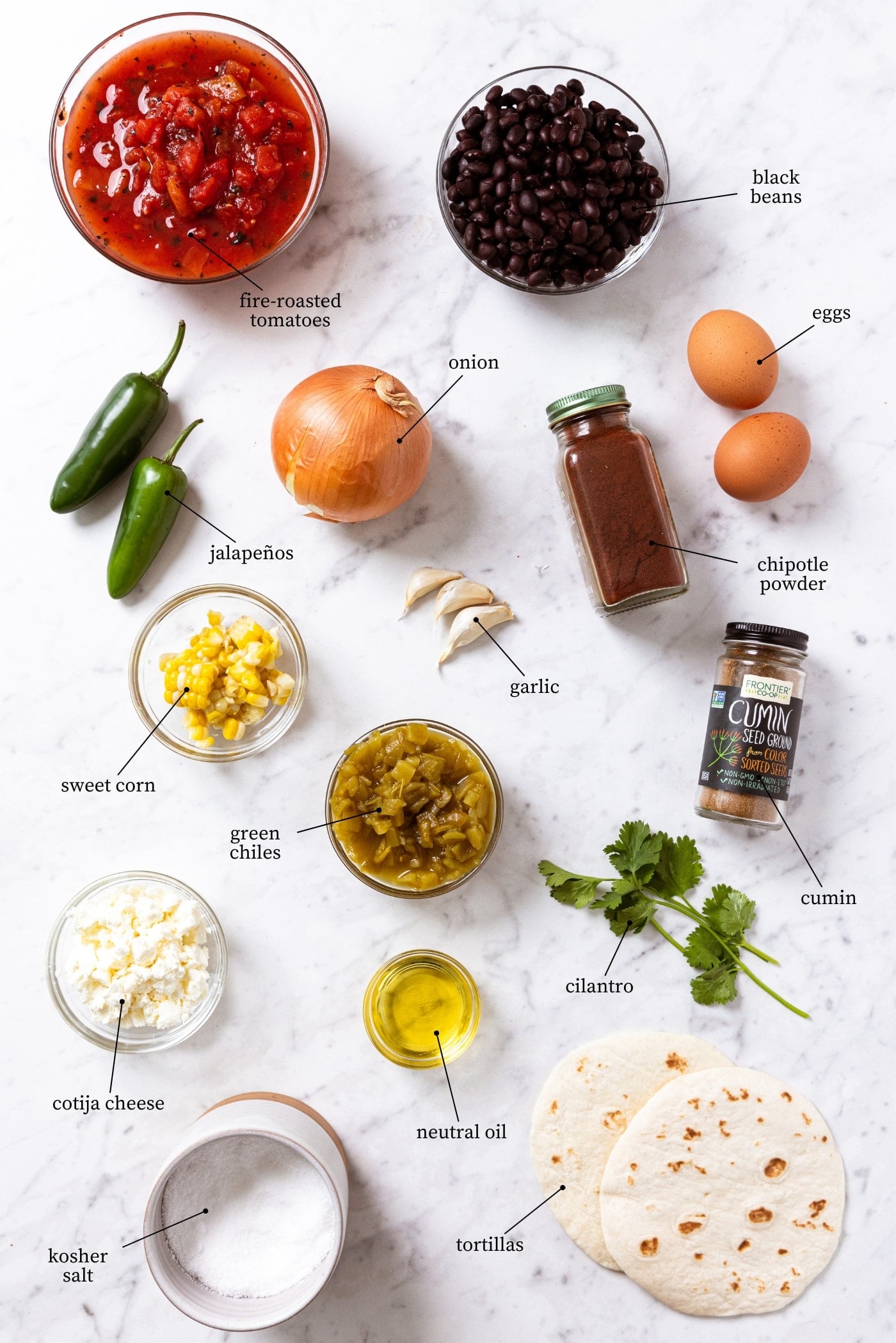 Marble surface filled with all ingredients needed to make a Tex-Mex spiced skillet dish including onion, jalapeños, black beans, tomatoes, tortillas, spices, eggs, and more.