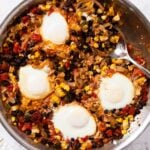 White marble surface with metal skillet filled with tomato, sweet corn, and black bean mixture with cooked soft eggs on top with spoon resting on the side of skillet.