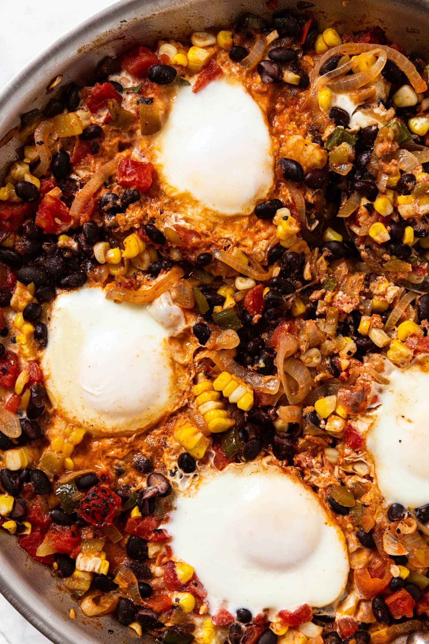 Metal skillet filled with cooked sweet corn, black beans, and tomato mixture topped with soft cooked eggs with spoon to the side.