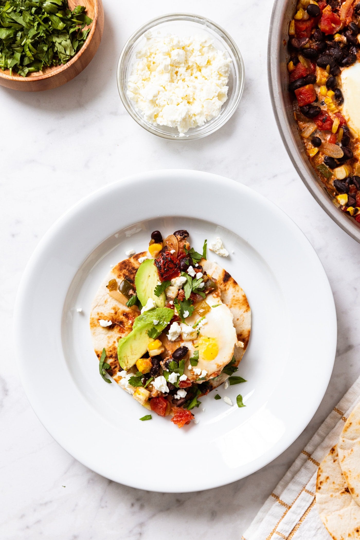 White plate filled with tortilla stuffed with black bean, corn, and tomato mixture and topped with cotija cheese, egg, and pieces of avocado all on white marble surface with cilantro and more cheese to the side.
