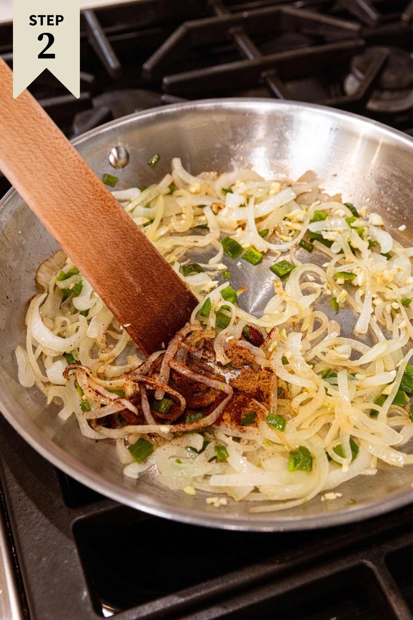 Metal skillet with diced jalapeños and sliced onions with a clump of brown spices before being mixed together with a wooden spatula.