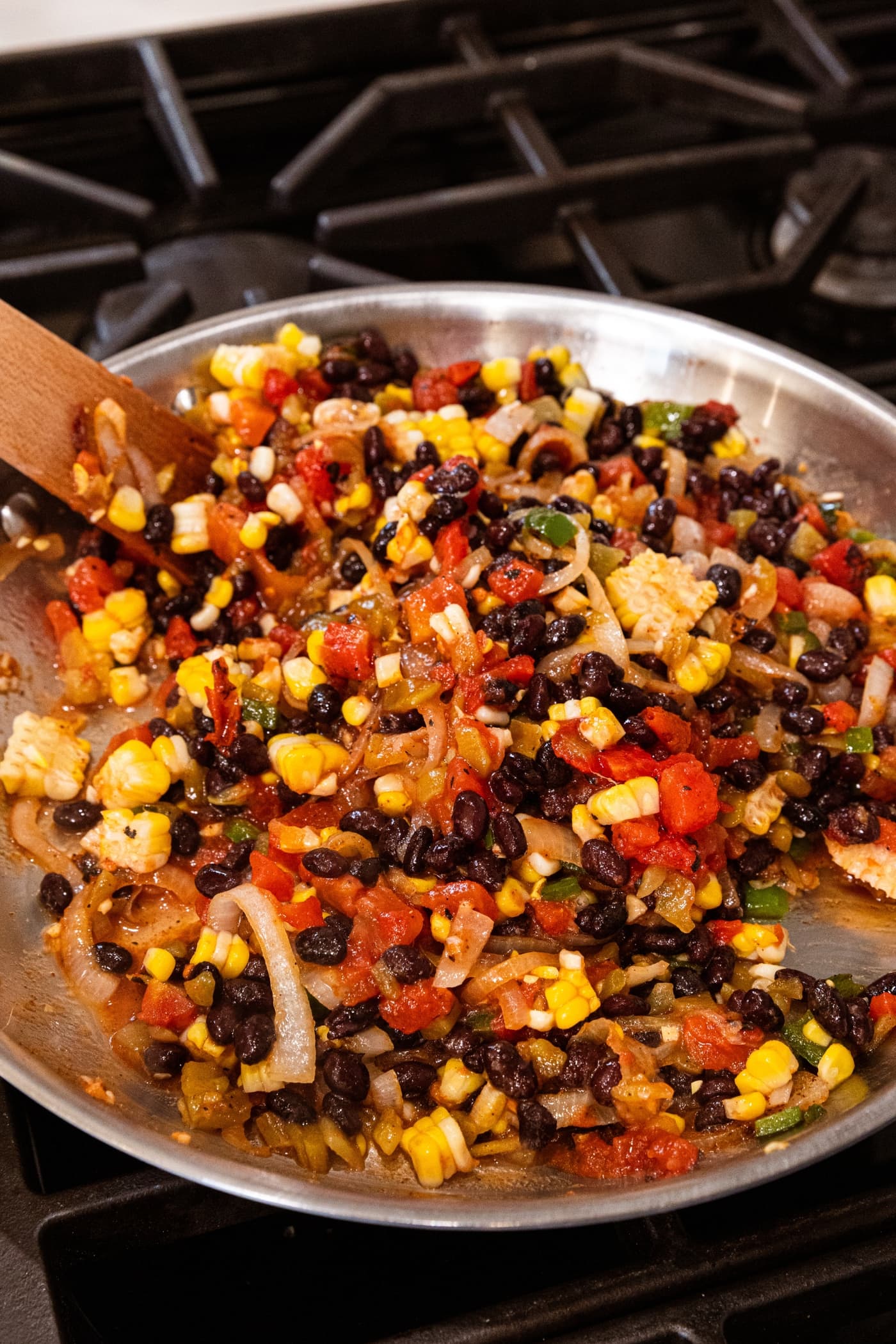 Black beans, corn, onions, and tomatoes all in metal skillet on stove being mixed together with wooden spatula.