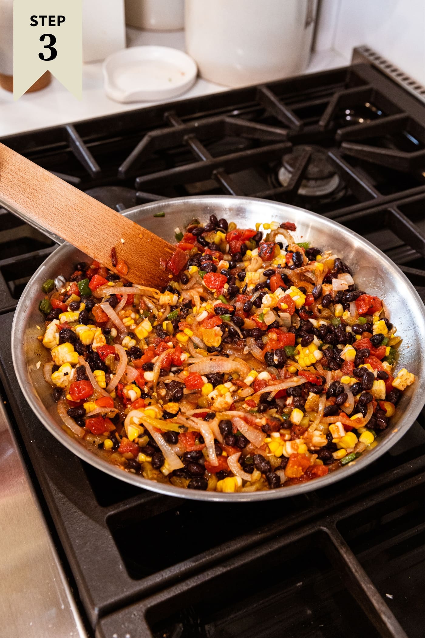Metal skillet filled with corn, black beans, and tomatoes on the stovetop being mixed together with wooden spatula.