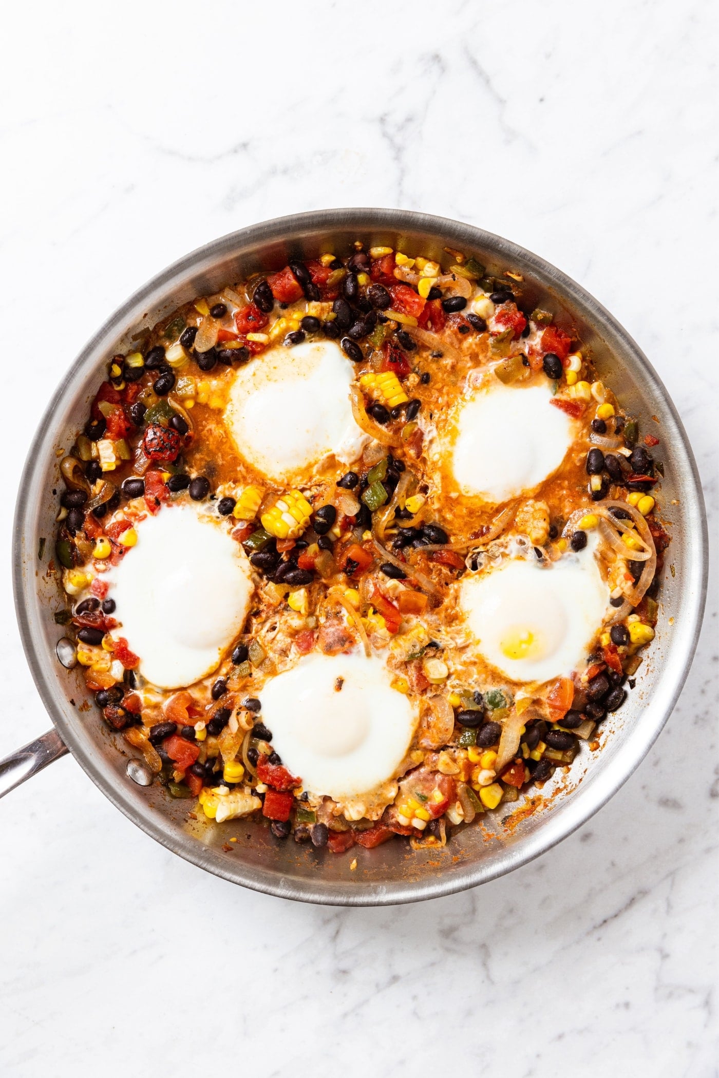 Metal skillet sitting on marble surface filled with corn, black bean, and tomato mixture with cooked soft eggs on top.
