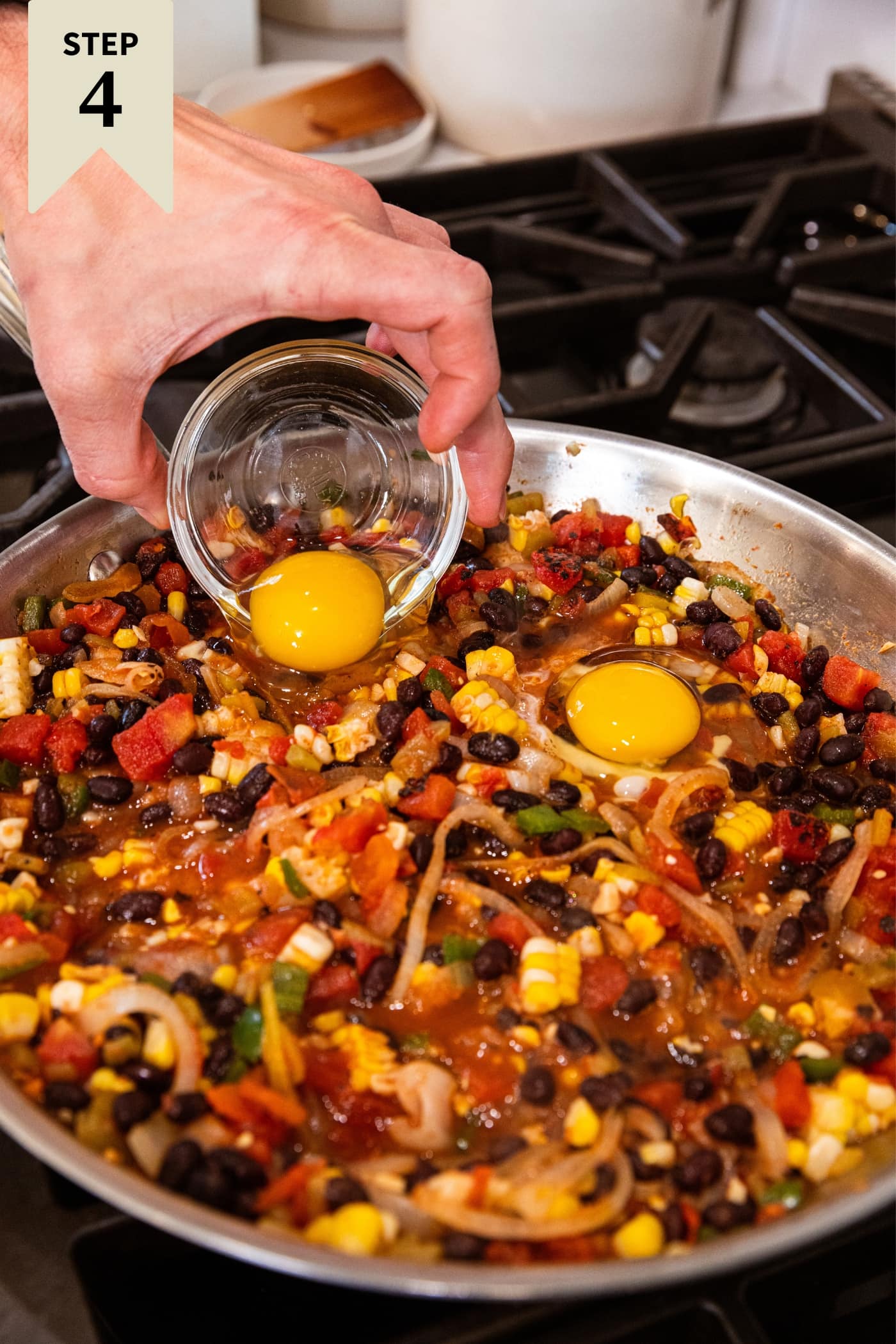 Hand holding small glass bowl with raw egg pouring it on top of mixture of beans, corn, and tomatoes in skillet.