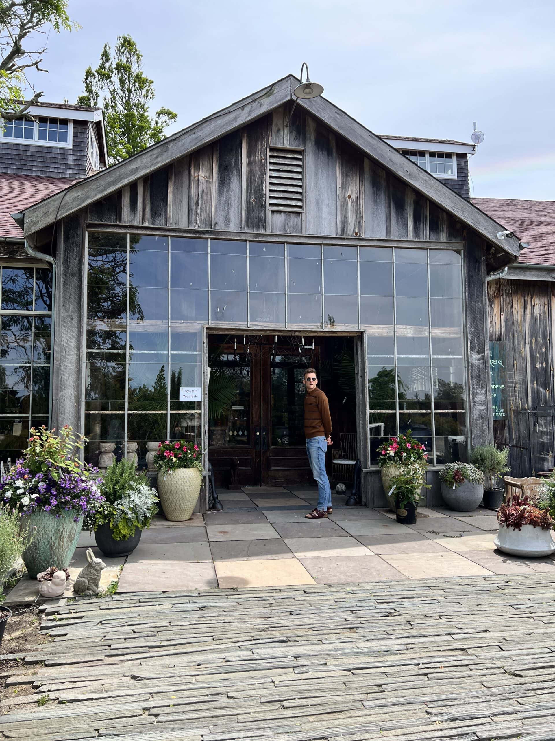 Kaleb Wyse wearing brown shirt standing at the entrance of a wood-clad landscaping store with urns filled with plants.