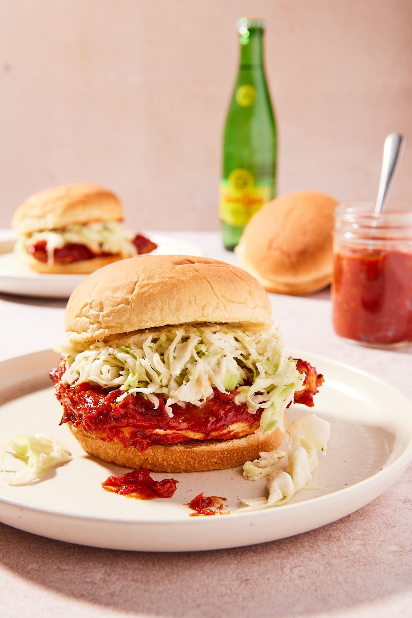 Plate sitting on light pink surface with chicken sandwich layered with coleslaw and more BBQ sauce with more buns and sauce in the background.