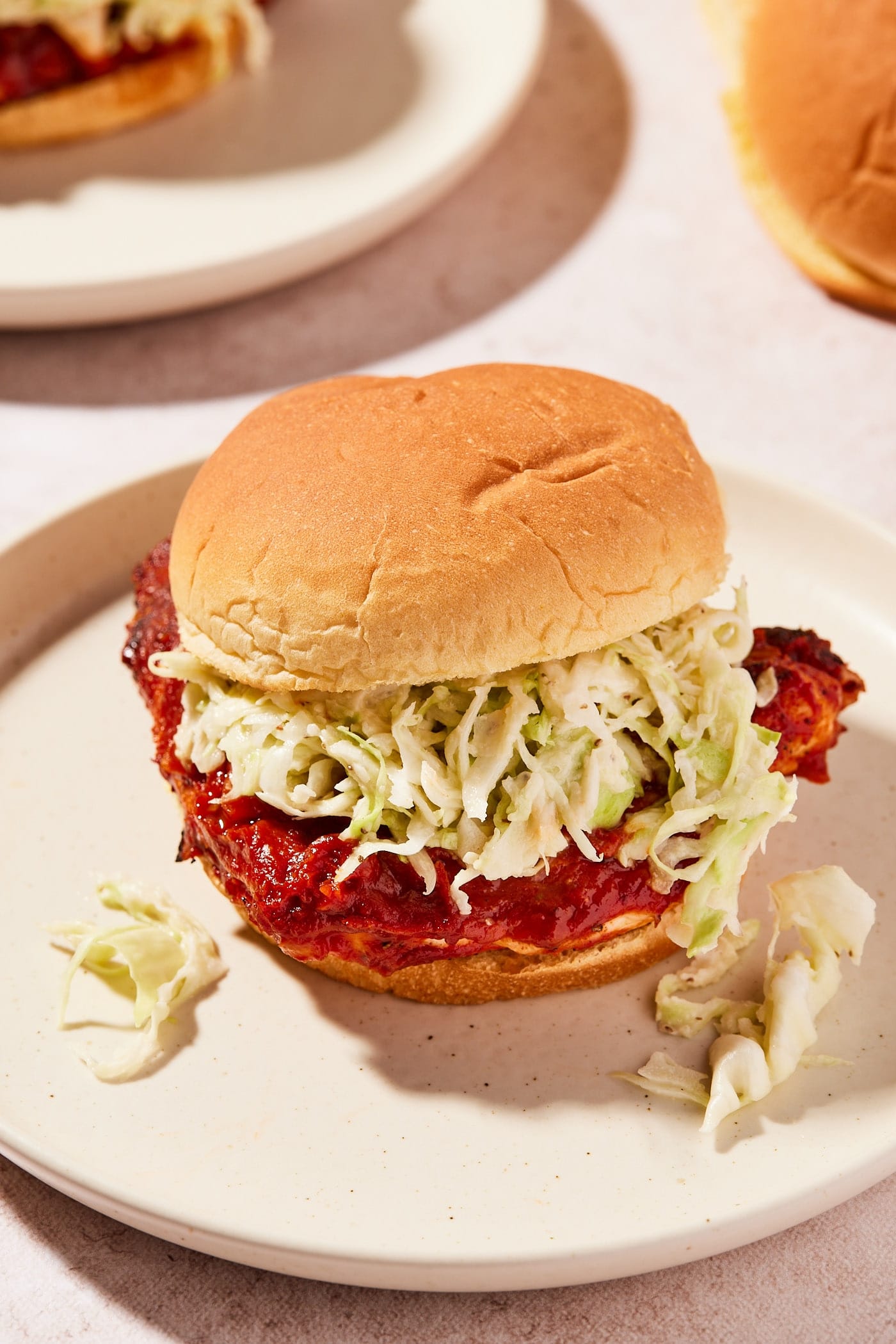 Tan-colored plate with BBQ chicken sandwich sitting with layer of coleslaw and some falling out with barbecue sauce all over.