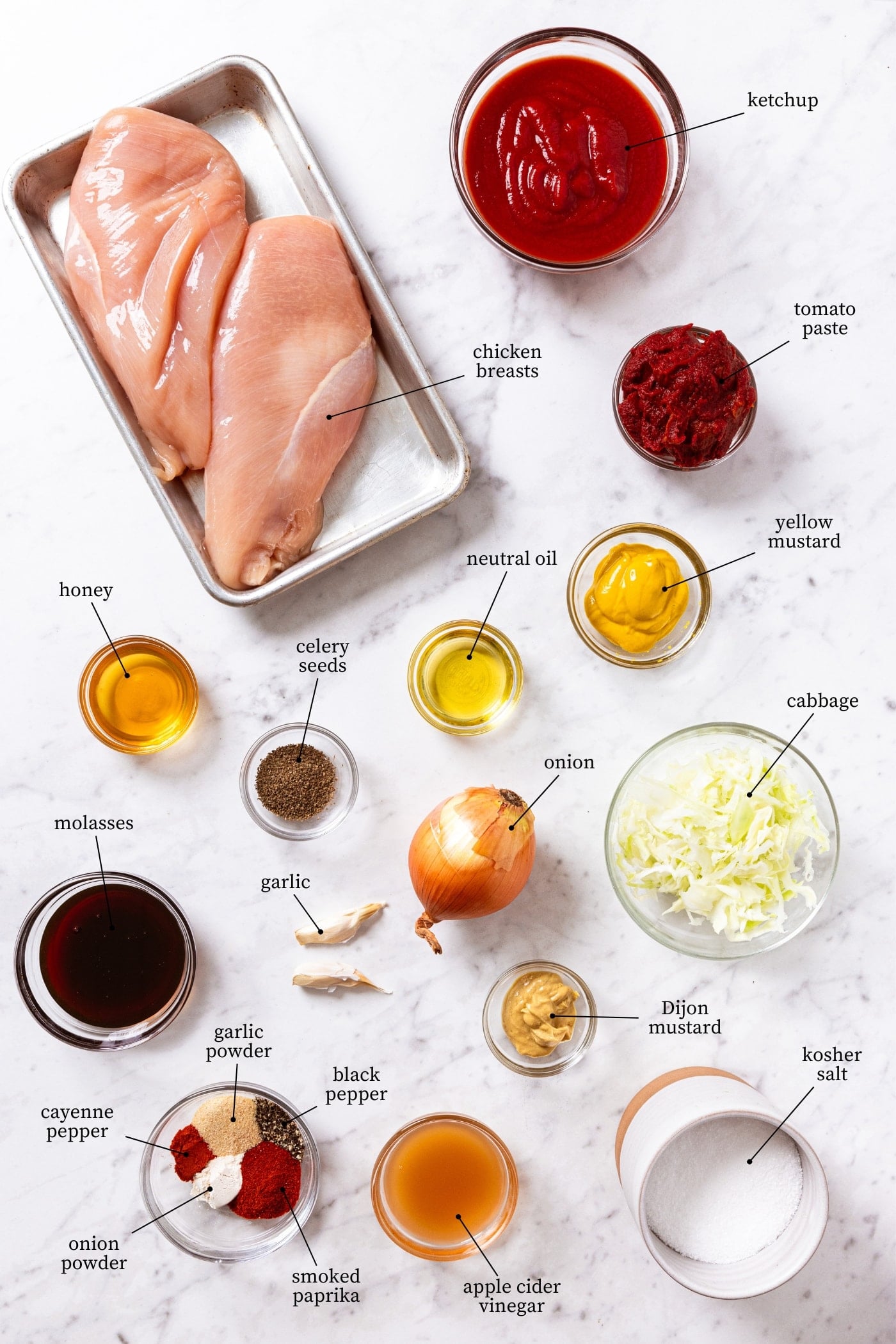 White marble surface filled with all ingredients needed to prepare a BBQ chicken sandwich including raw chicken breasts, tomato paste, spices, molasses, onion, cabbage, yellow mustard, and more.