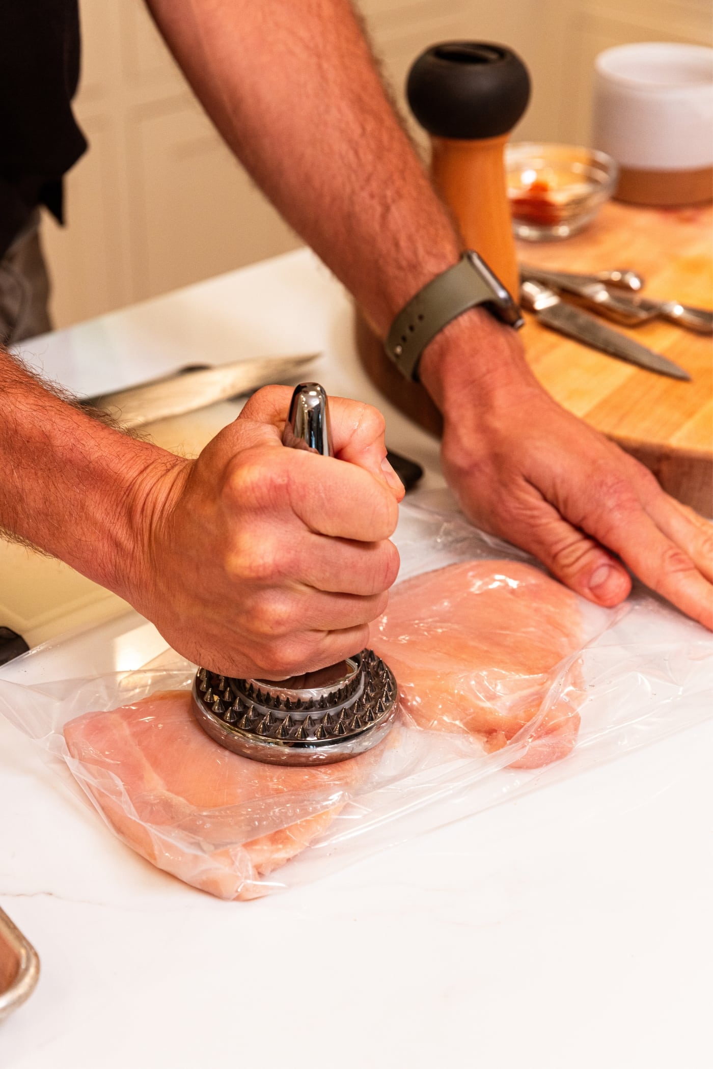 Hand holding meat pounder pounding raw chicken breasts in plastic bag on white surface.